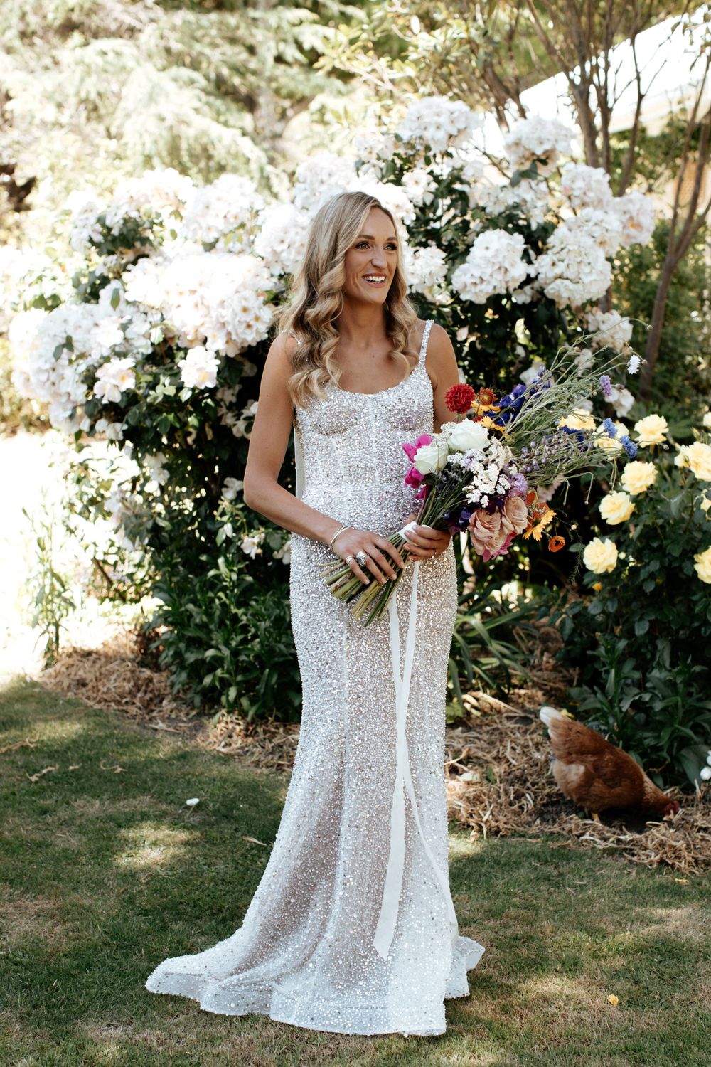 Bride in a sequined wedding gown poses in front of white floral garden backdrop.