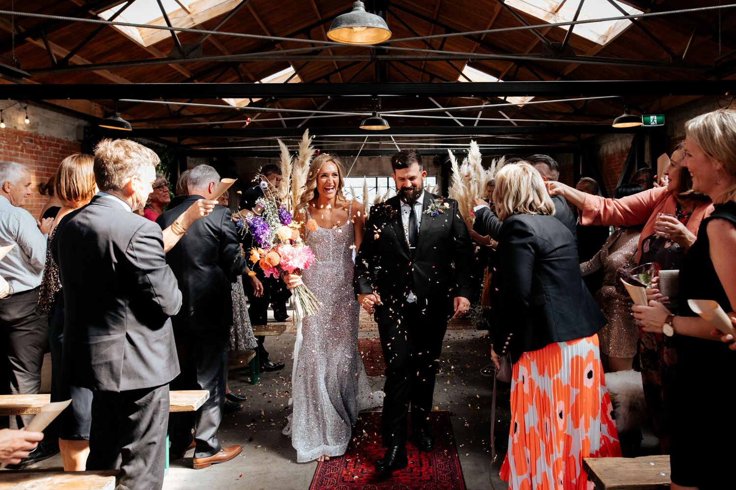 Wedding recessional with guests throwing confetti in rustic brick venue with overhead lighting.