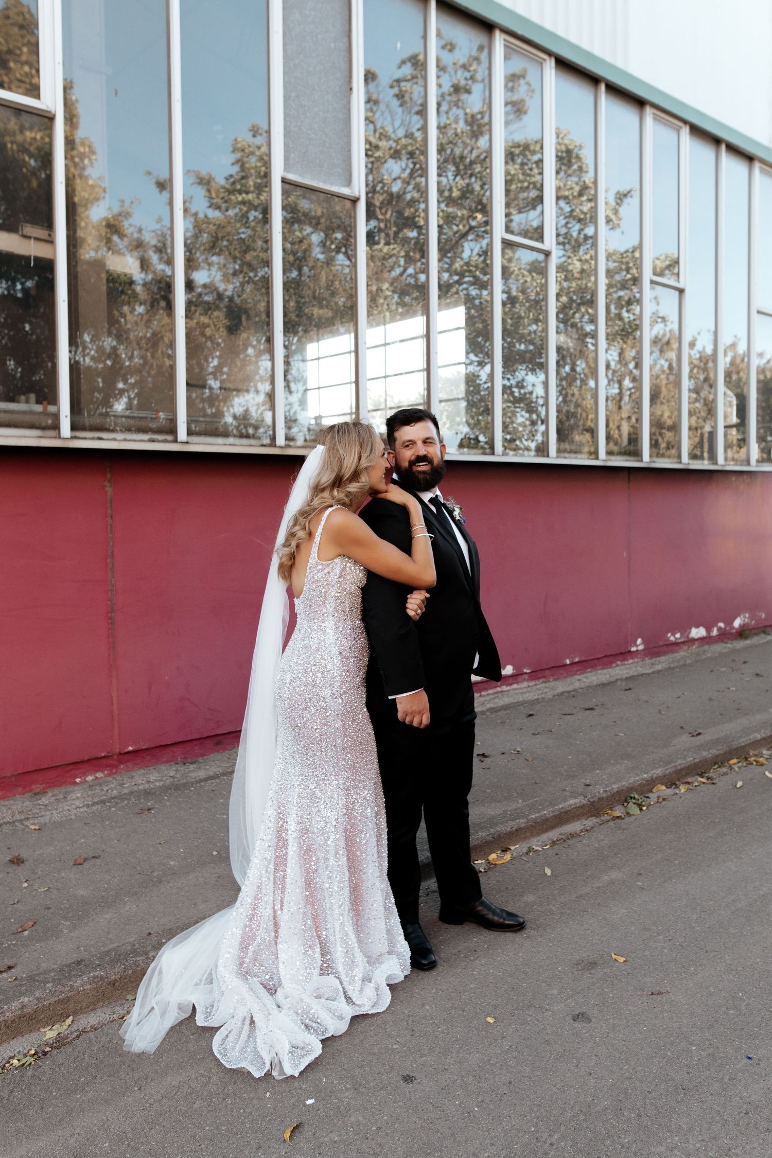 A couple embraces in front of a red wall with large windows during their wedding photoshoot.