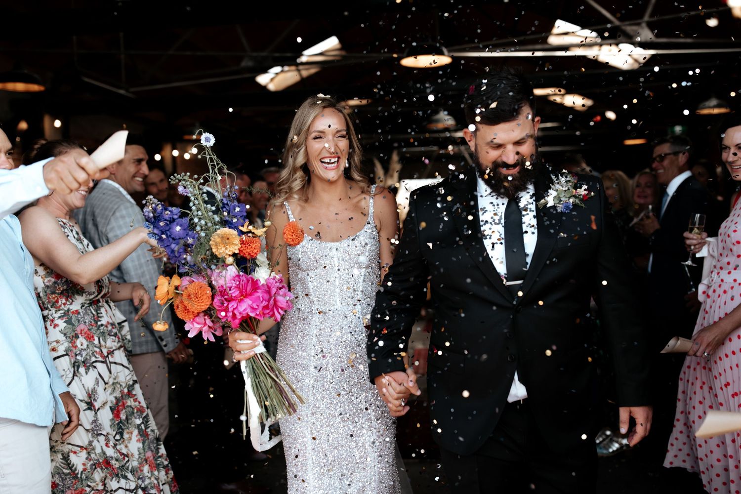 Newlyweds walk through shower of confetti holding colorful bouquet in industrial wedding venue.