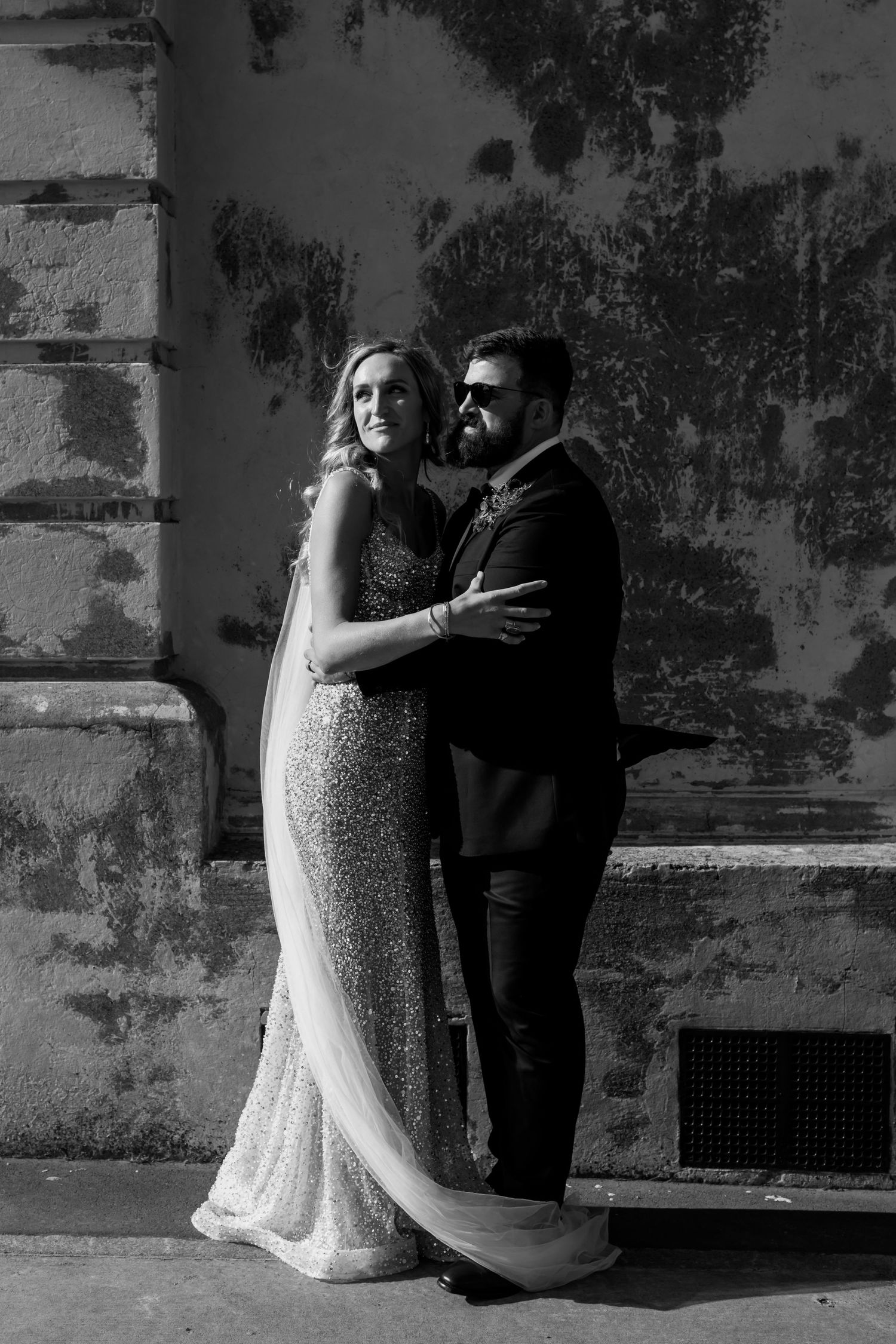 A black and white portrait of newlyweds posing against a weathered wall.