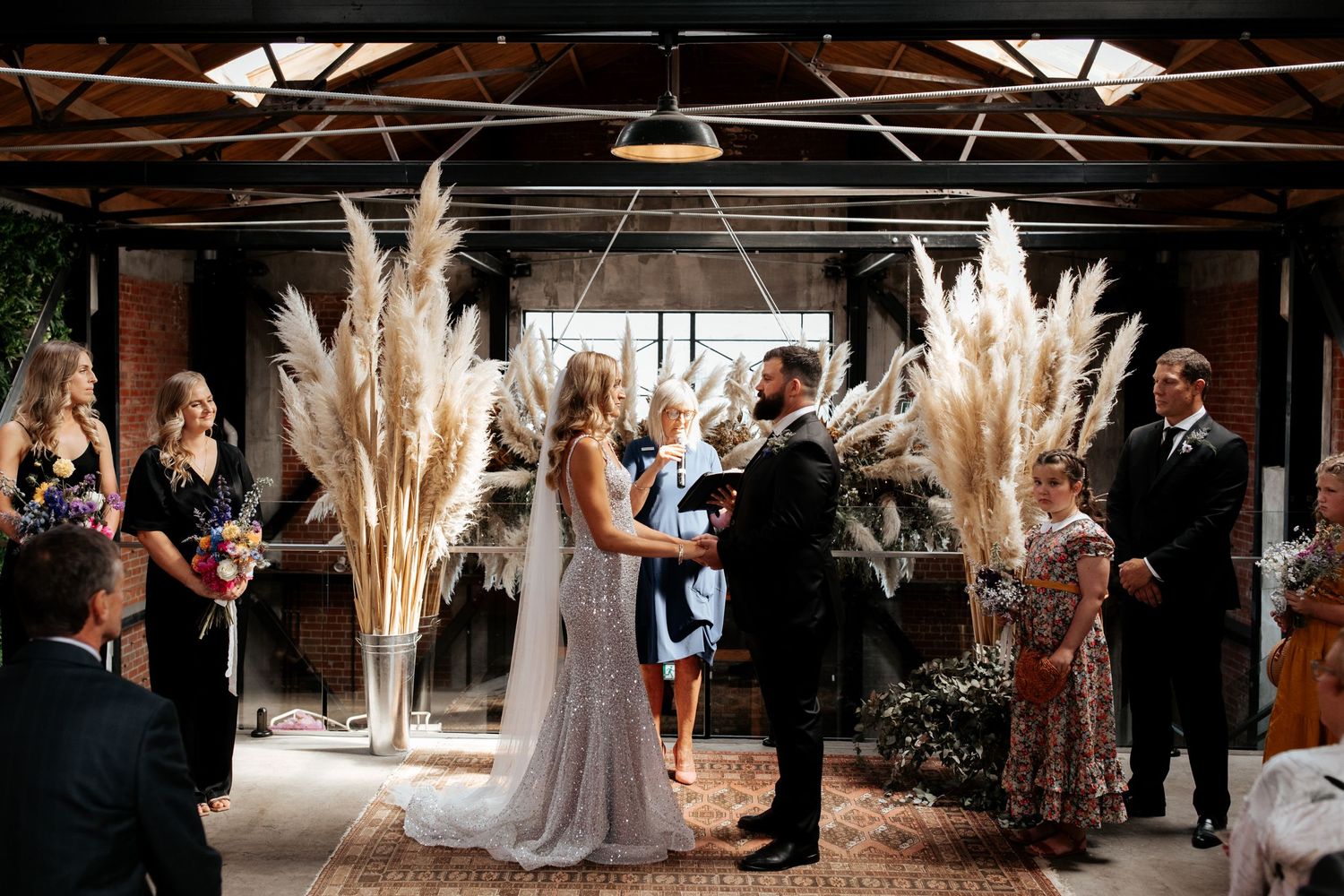 Wedding ceremony in industrial warehouse venue with dramatic pampas grass arrangements and exposed beams.