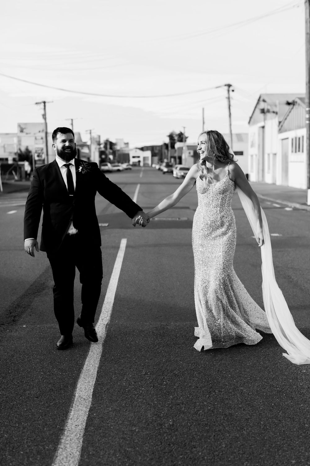 Black and white image of couple holding hands while walking down center of empty street.