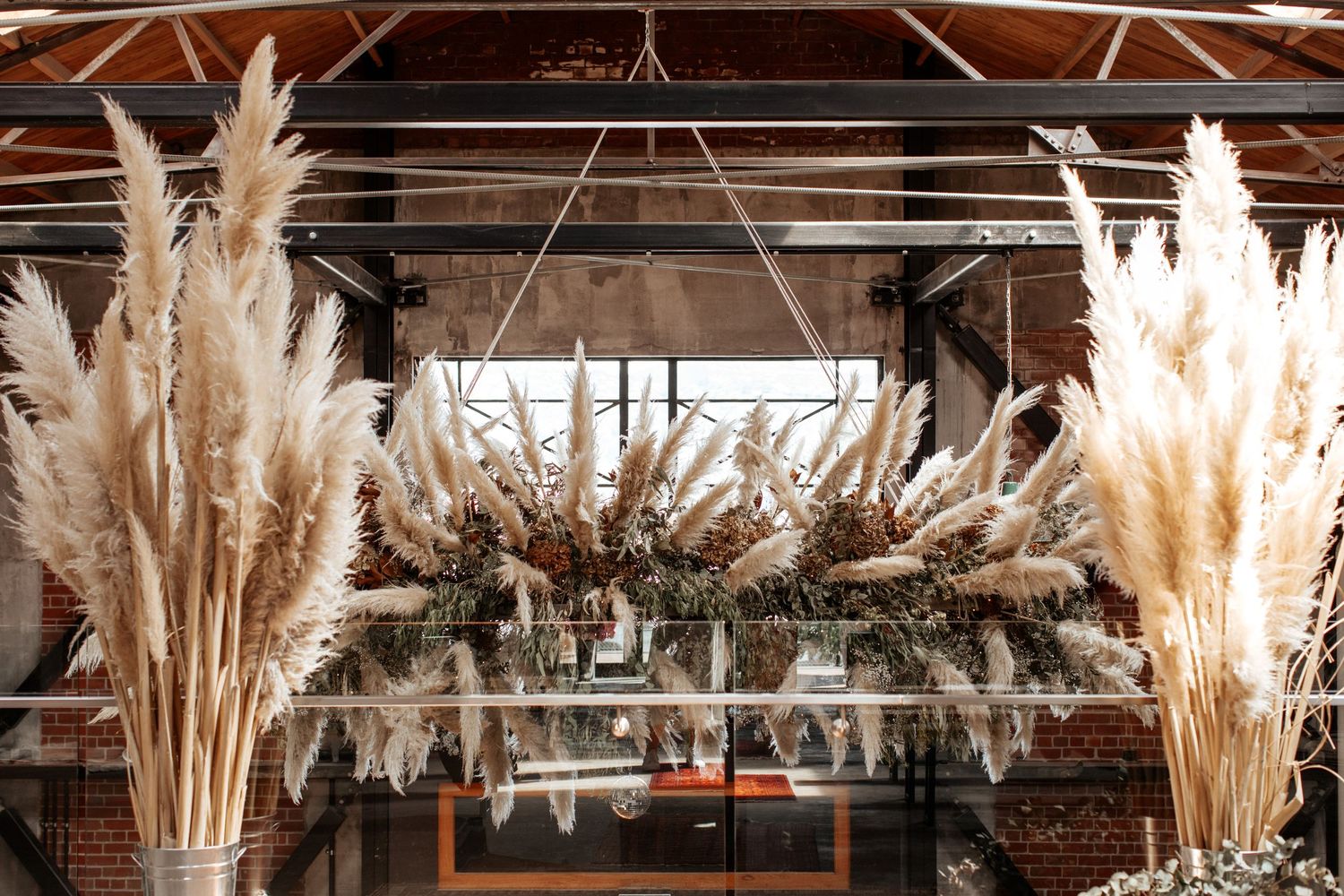 Decorative dried pampas grass and floral arrangements hanging from wooden beams in an indoor venue.