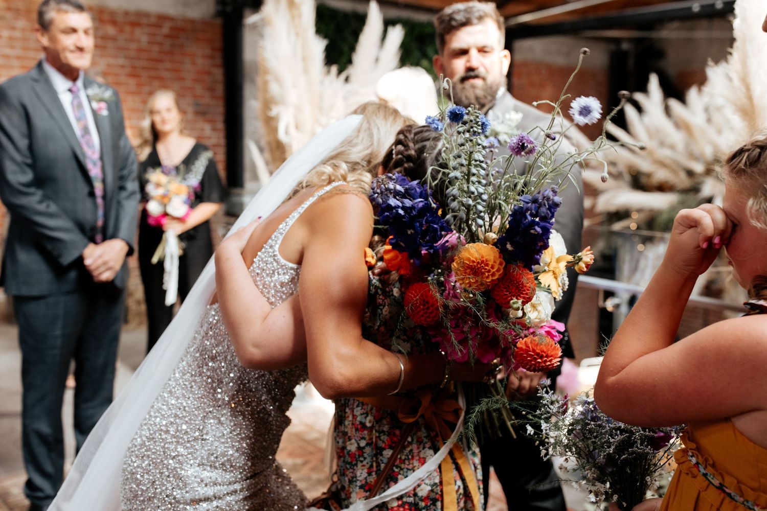Confetti moment during wedding ceremony in rustic brick venue featuring vibrant florals and pampas grass.