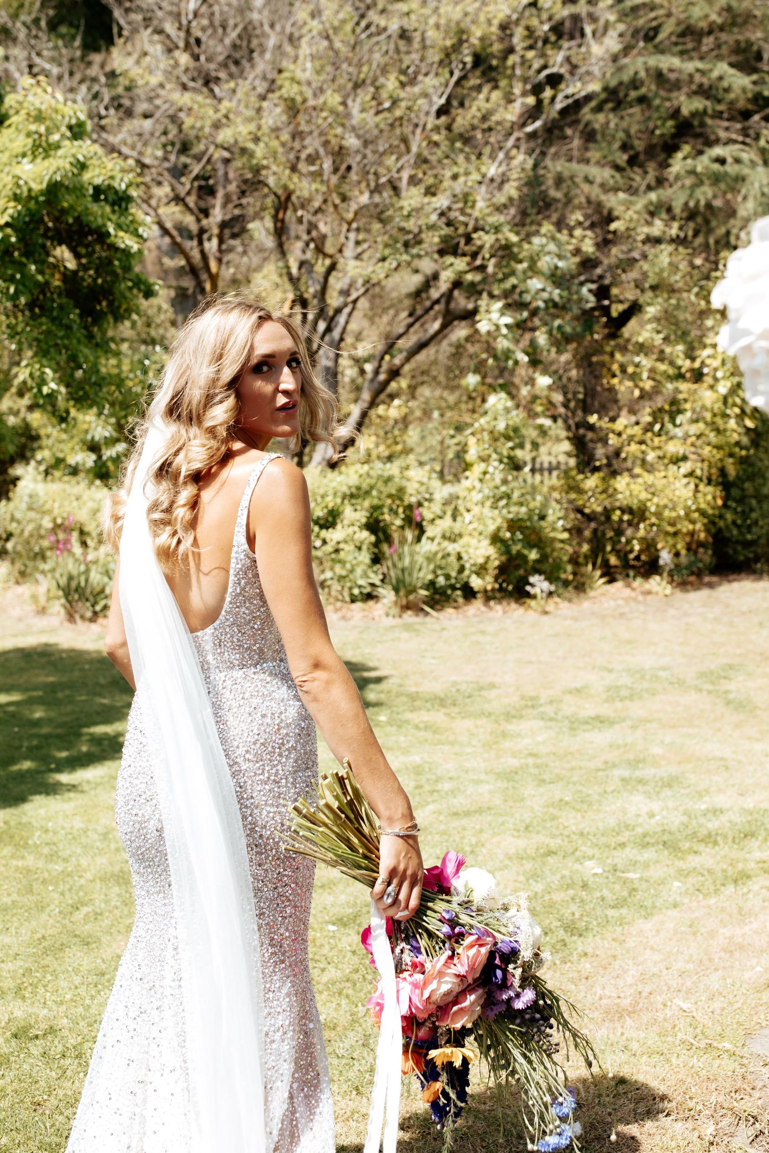 Side view of sparkly wedding dress and colorful bouquet in sunny garden setting.