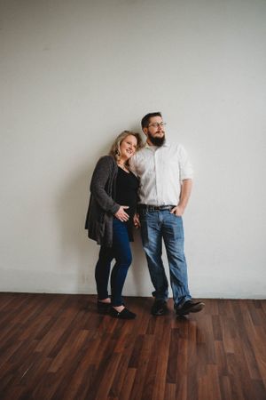 Casual portrait of couple standing against white wall with hardwood floors.