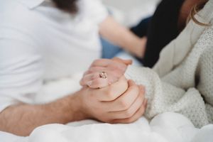 Close-up shot of hands clasped together showing engagement rings on white bedding.