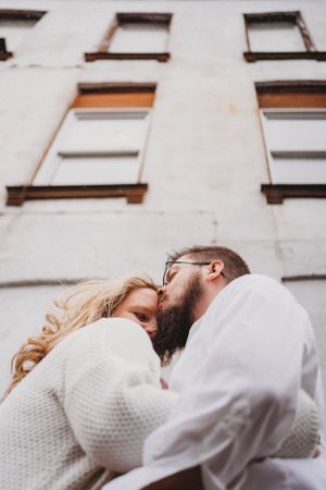 Couple embracing against an urban white building facade with vintage windows.