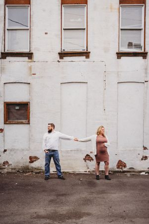 Playful couple holding hands at arms length against weathered white building wall.