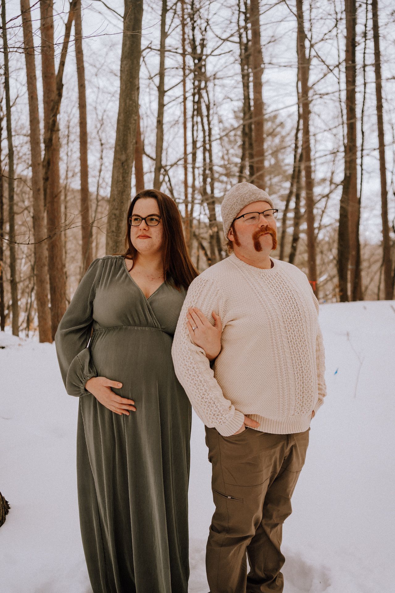 Couple poses for winter maternity photo in snowy forest setting.