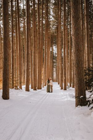 Romantic winter scene of couple standing among tall pine trees on snow-covered path.
