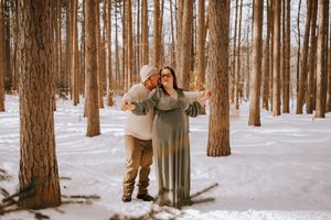 A couple embracing in a snowy forest with tall pine trees surrounding them.