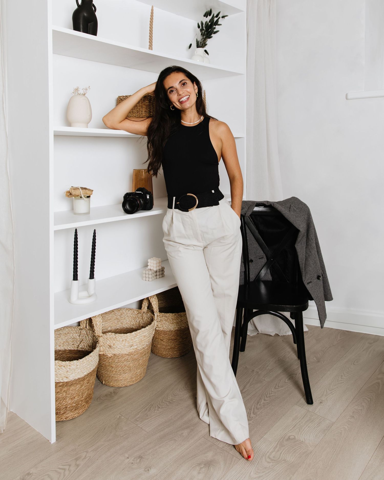 Woman in casual black tank top and white pants posing against white shelving with home decor.