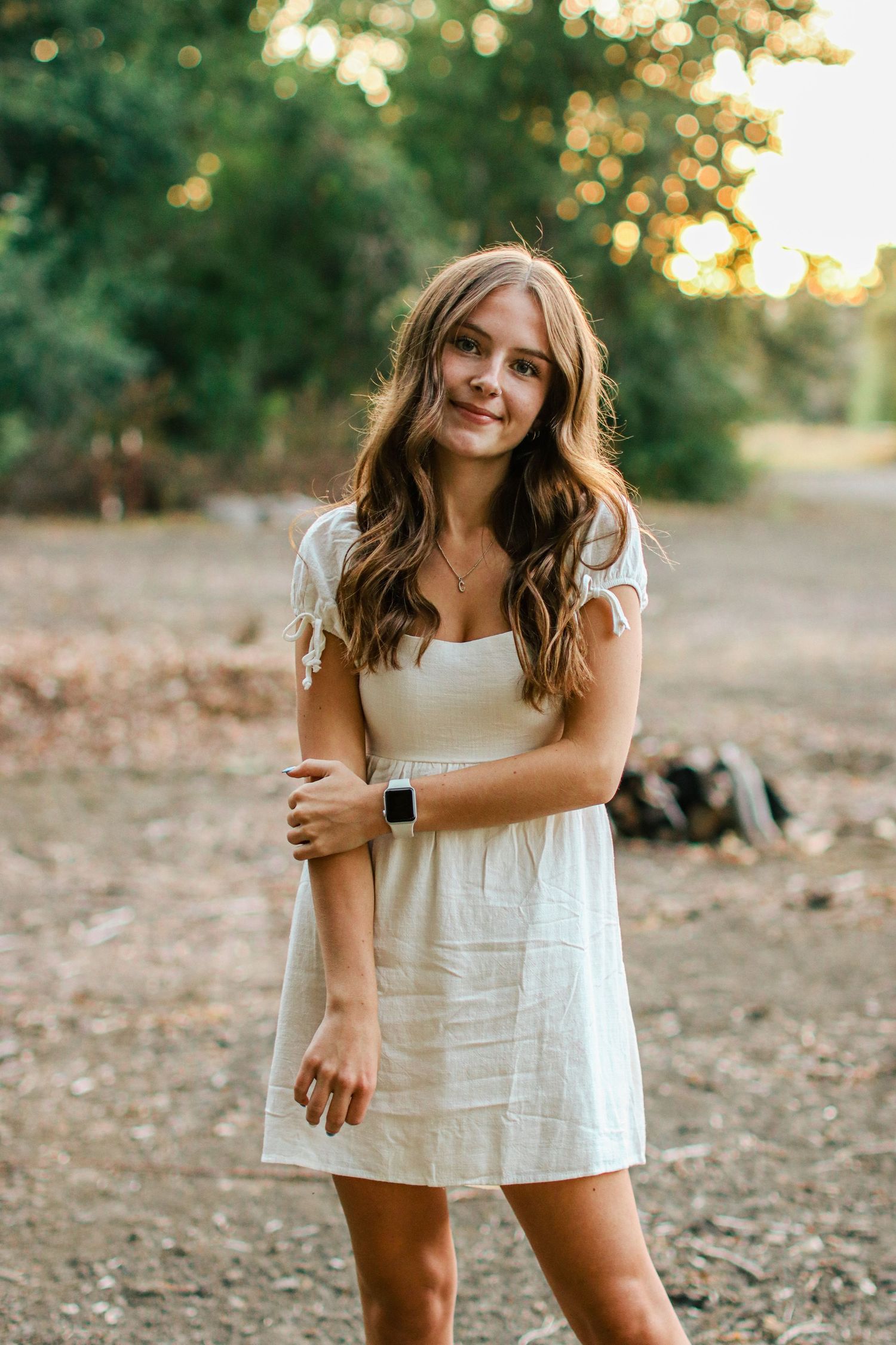 Young woman in white mini dress standing outdoors at golden hour with long wavy brown hair.