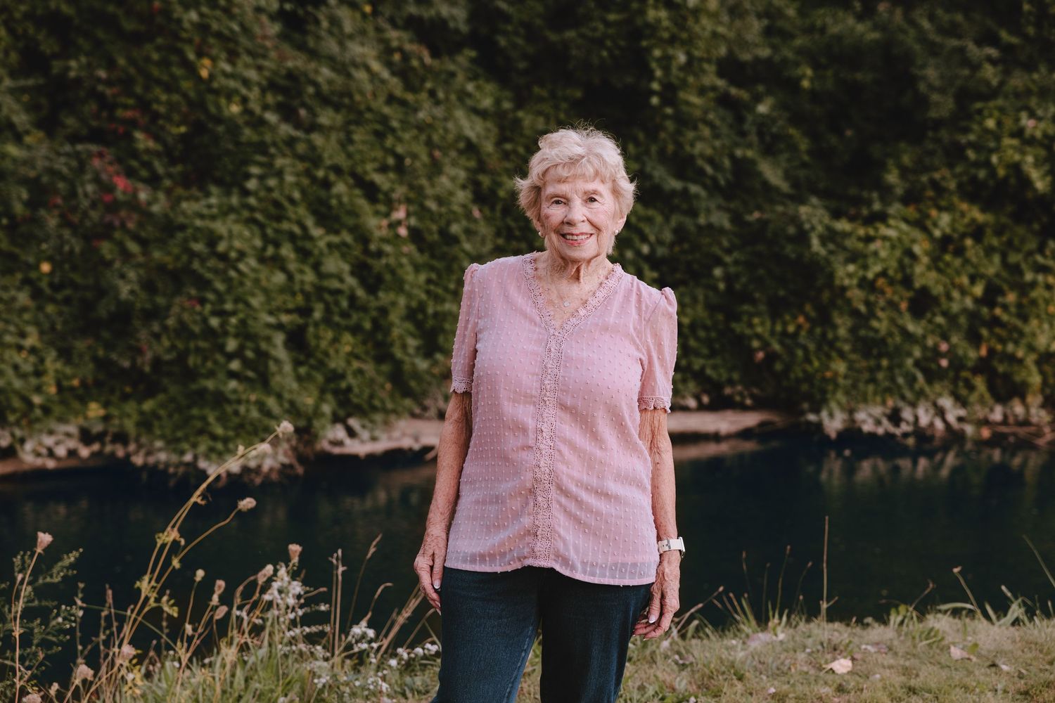 Woman in pink sleeveless top smiling outdoors against green foliage background.