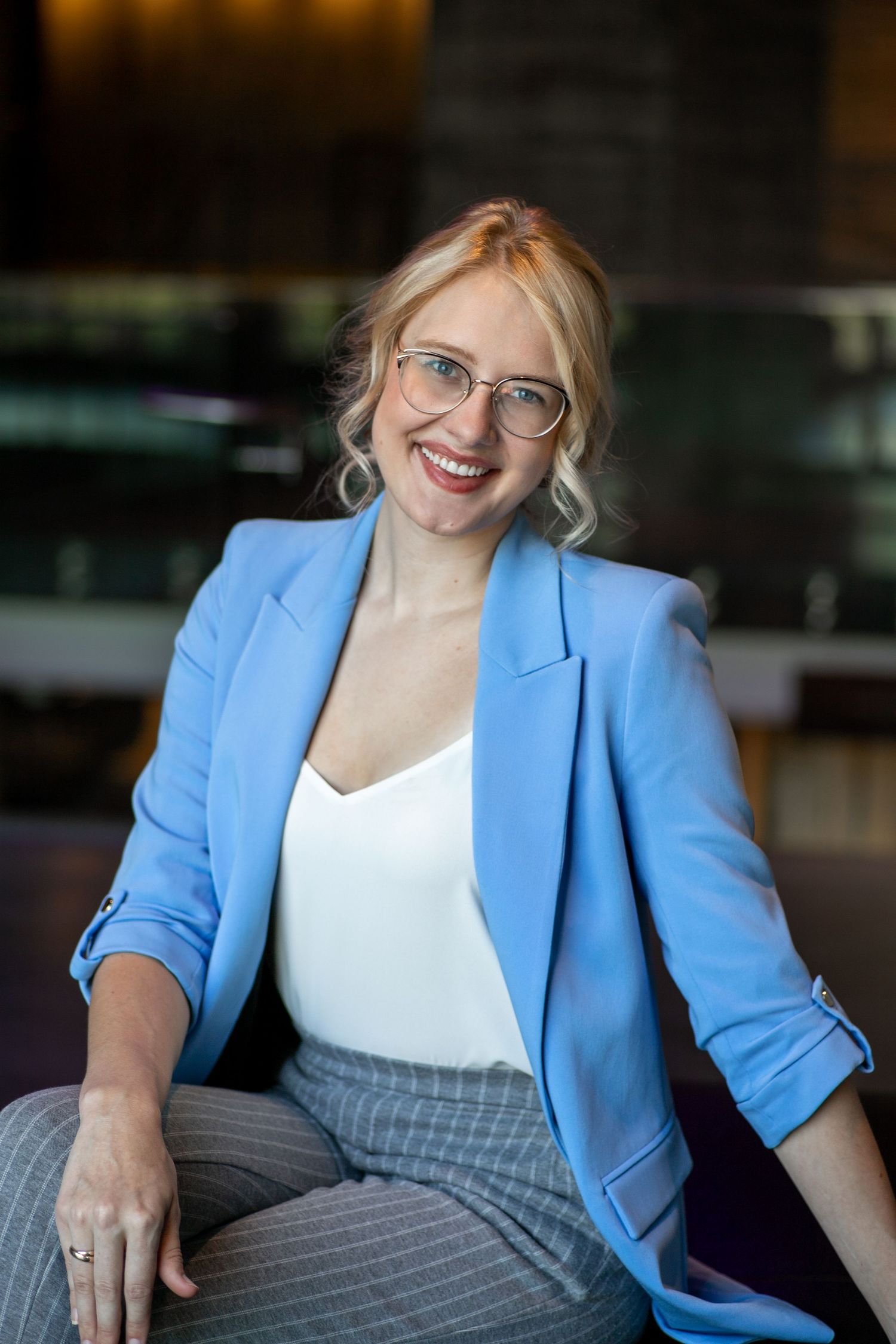 Professional headshot of a young white woman with blonde curled hair in a light blue blazer and glasses seated indoors.