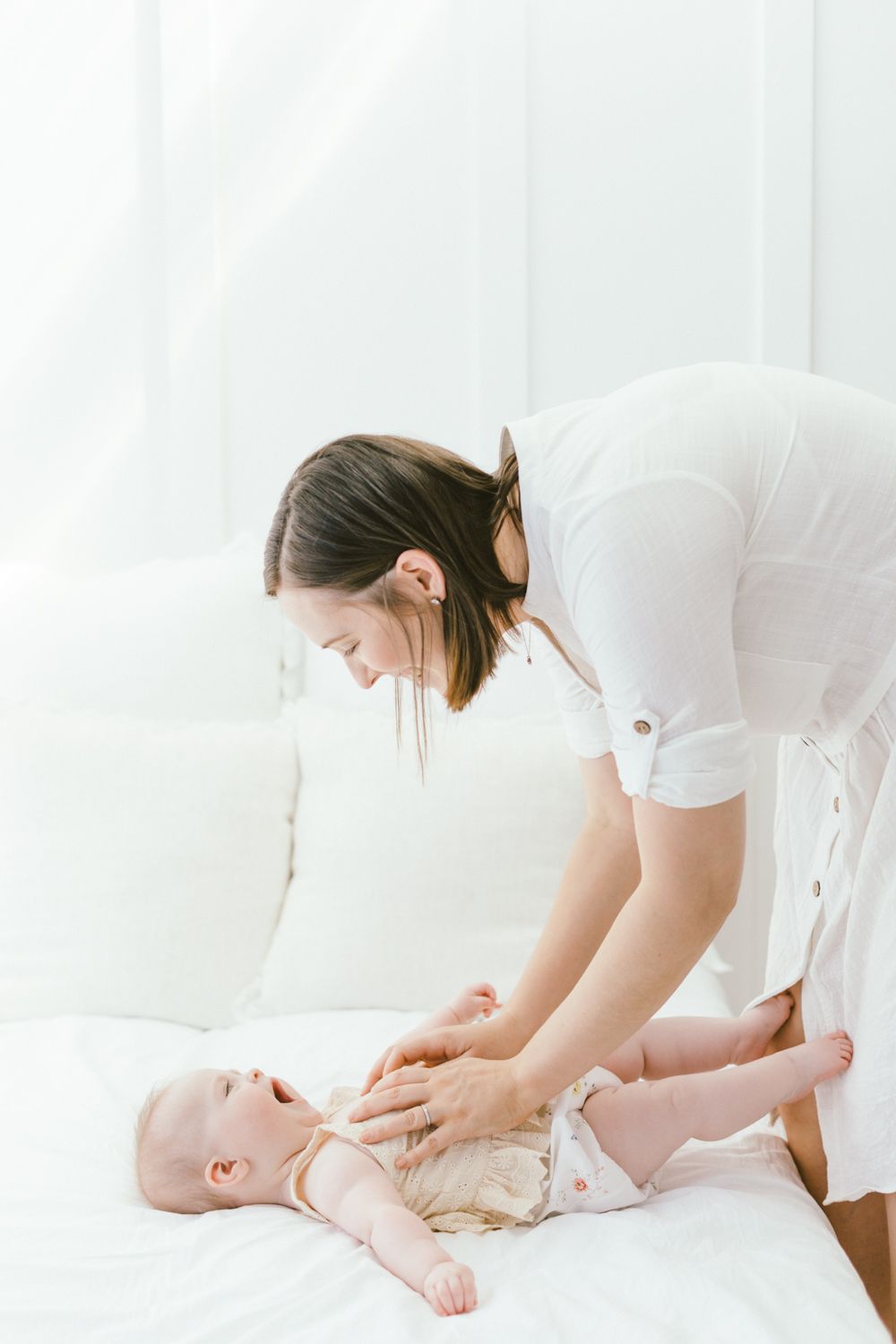 Woman in white shirt leaning over baby on white couch during playtime.
