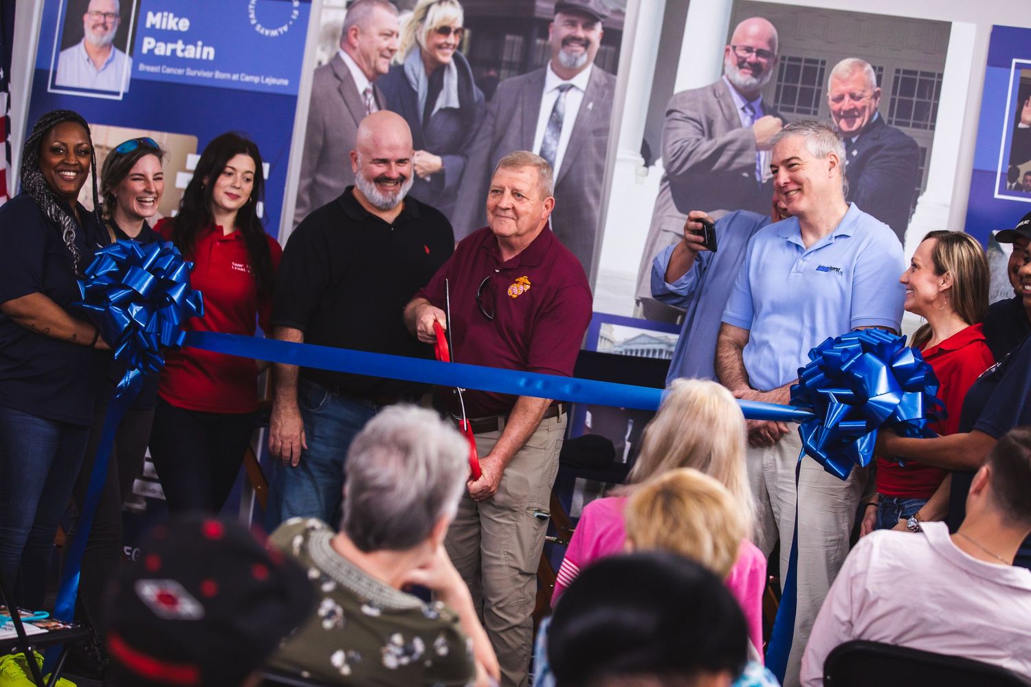 Crowd gathering at ribbon cutting ceremony with blue ribbon and decorative bows.