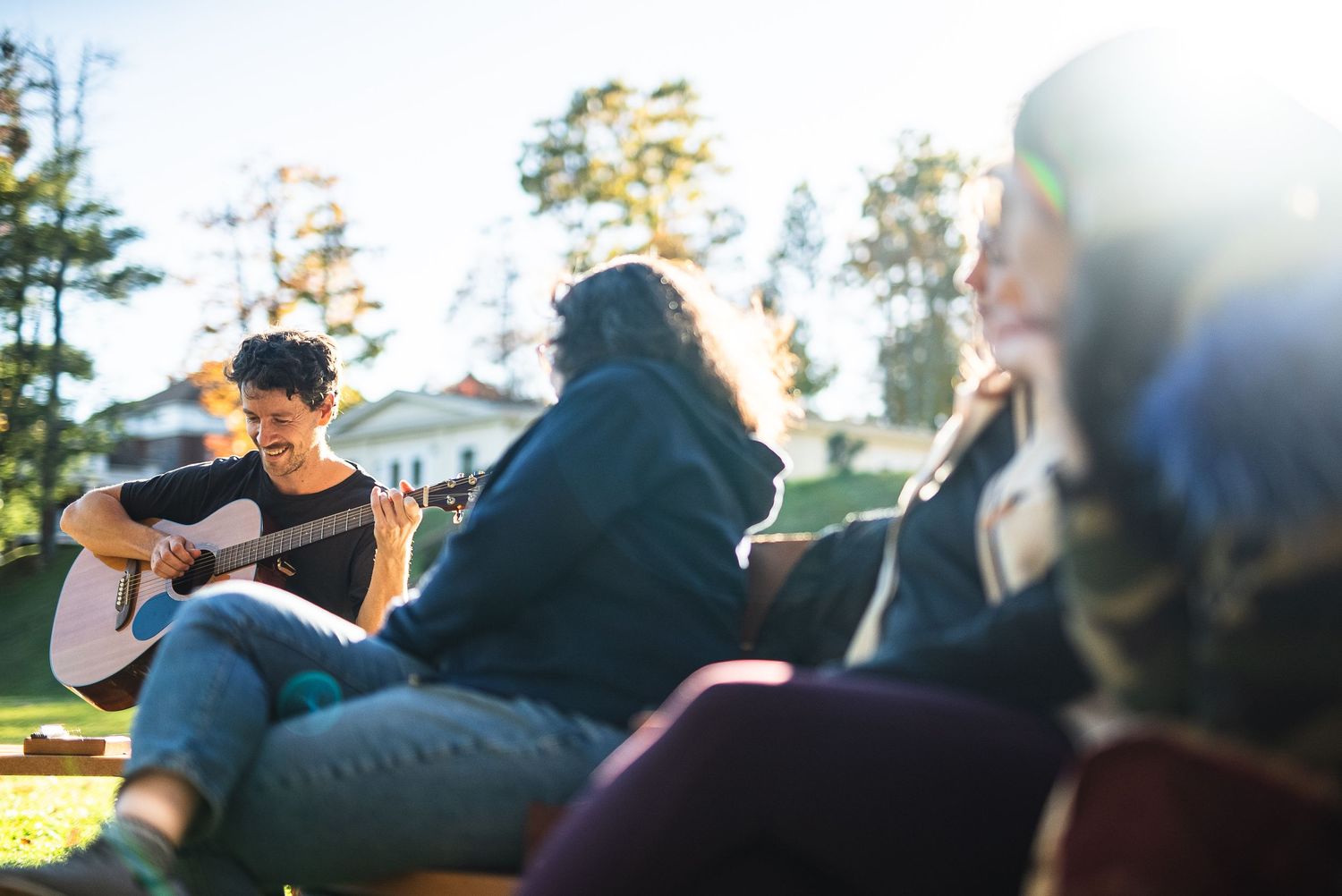 A group enjoys an outdoor acoustic guitar session in a sunny park setting.