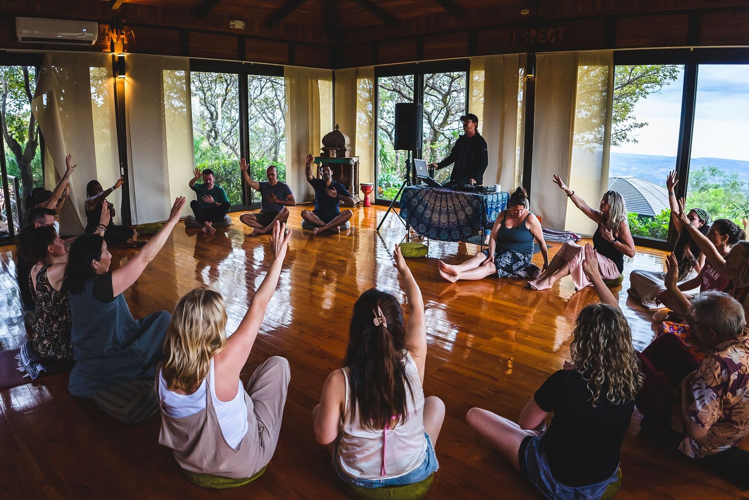 Group meditation session in a sunlit wooden floor room with panoramic views.
