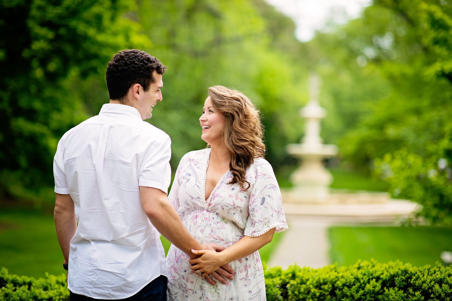 Couple in white attire share an intimate moment in a garden with fountain backdrop.