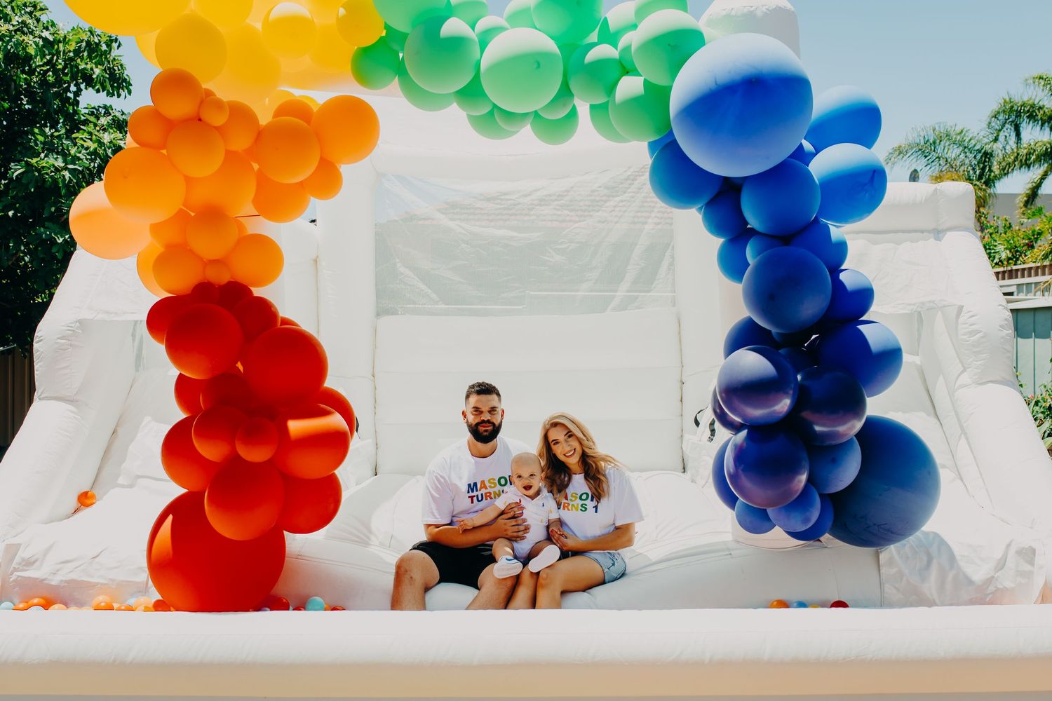 Rainbow balloon arch installation with white backdrop and seating area.