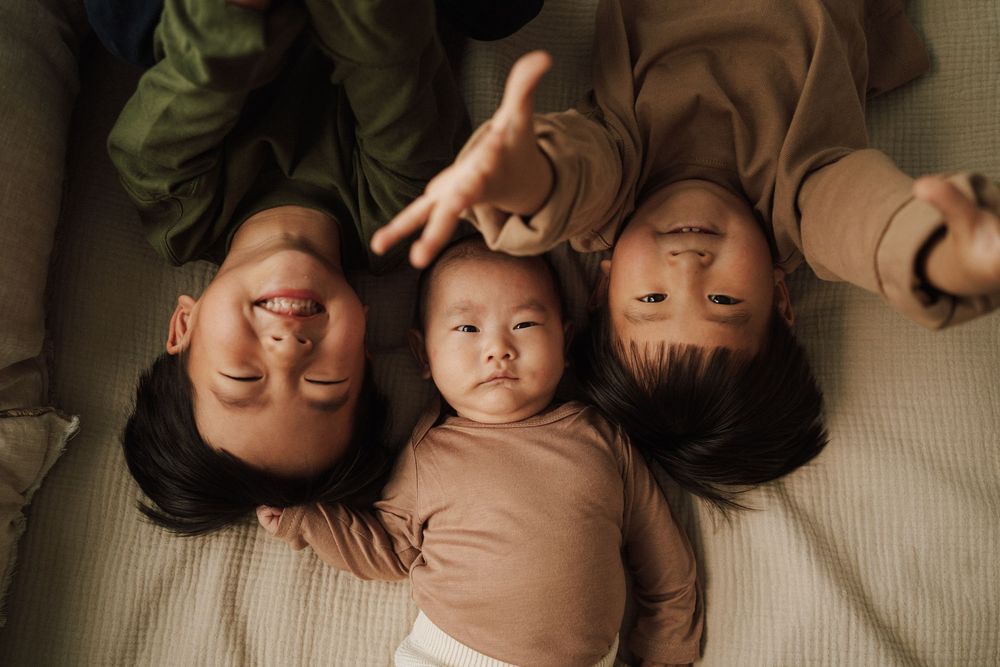 Three children lying together on a bed, two older kids framing a calm baby in the center, viewed from above.