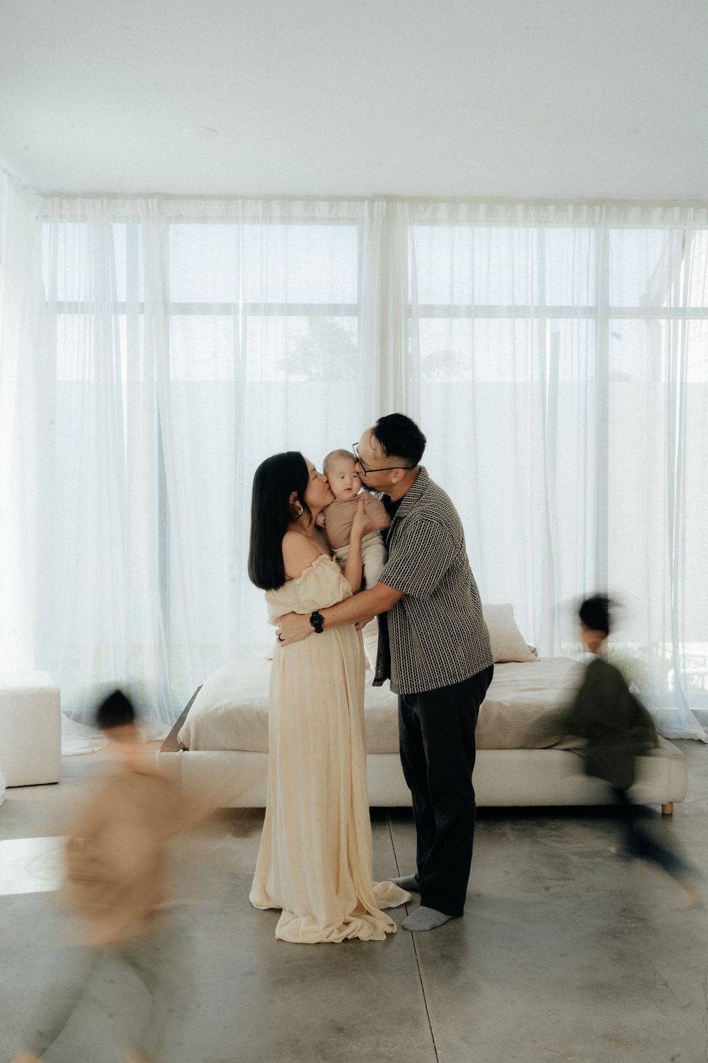 Parents kissing baby in bright white room while children run past in motion blur, modern minimalist family portrait.