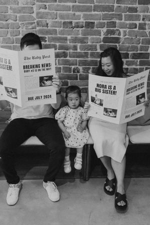 People sitting against brick wall reading newspapers with creative pregnancy announcement.