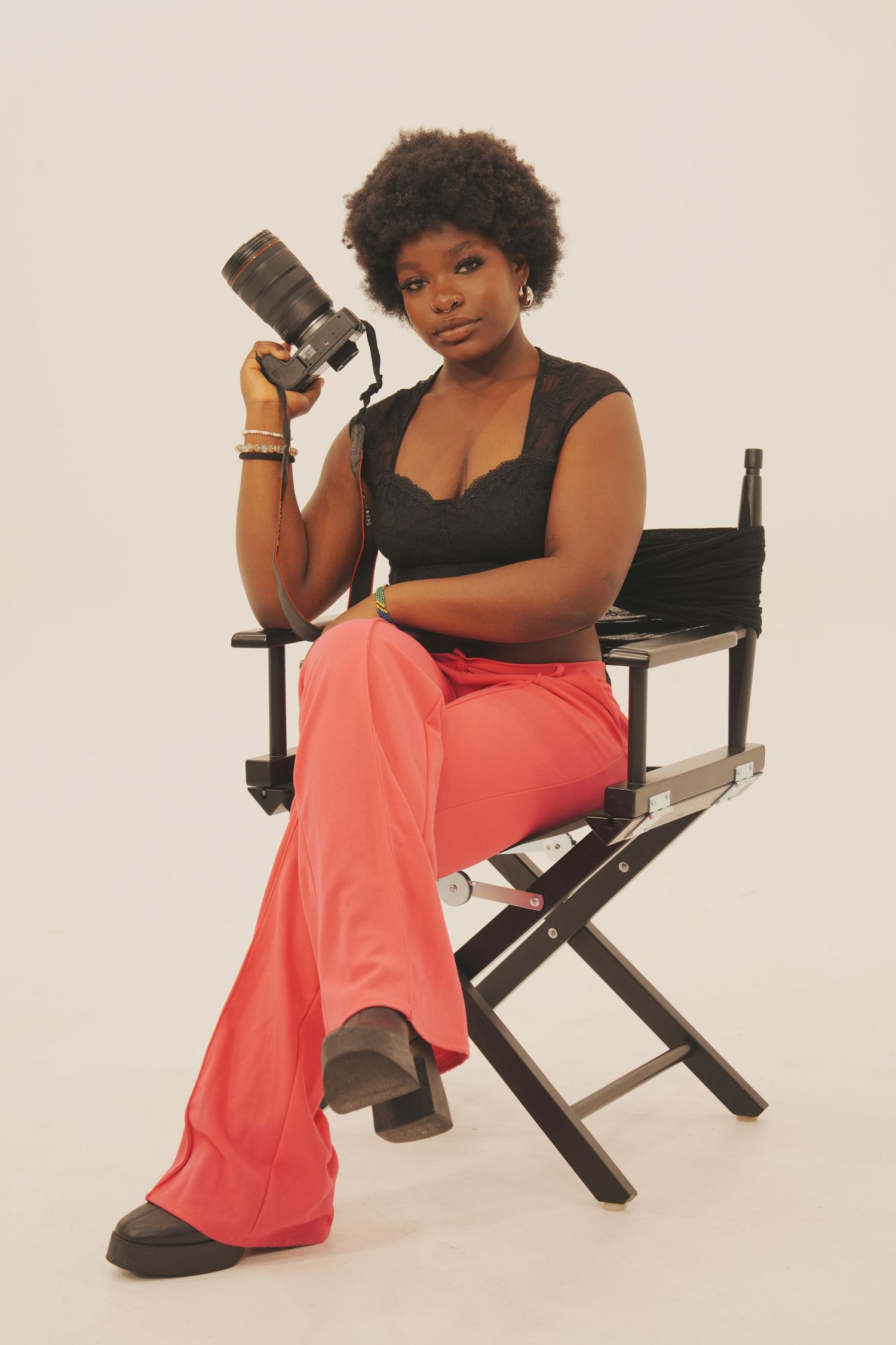 Professional photographer sits in director's chair wearing black top and coral pants while holding camera.