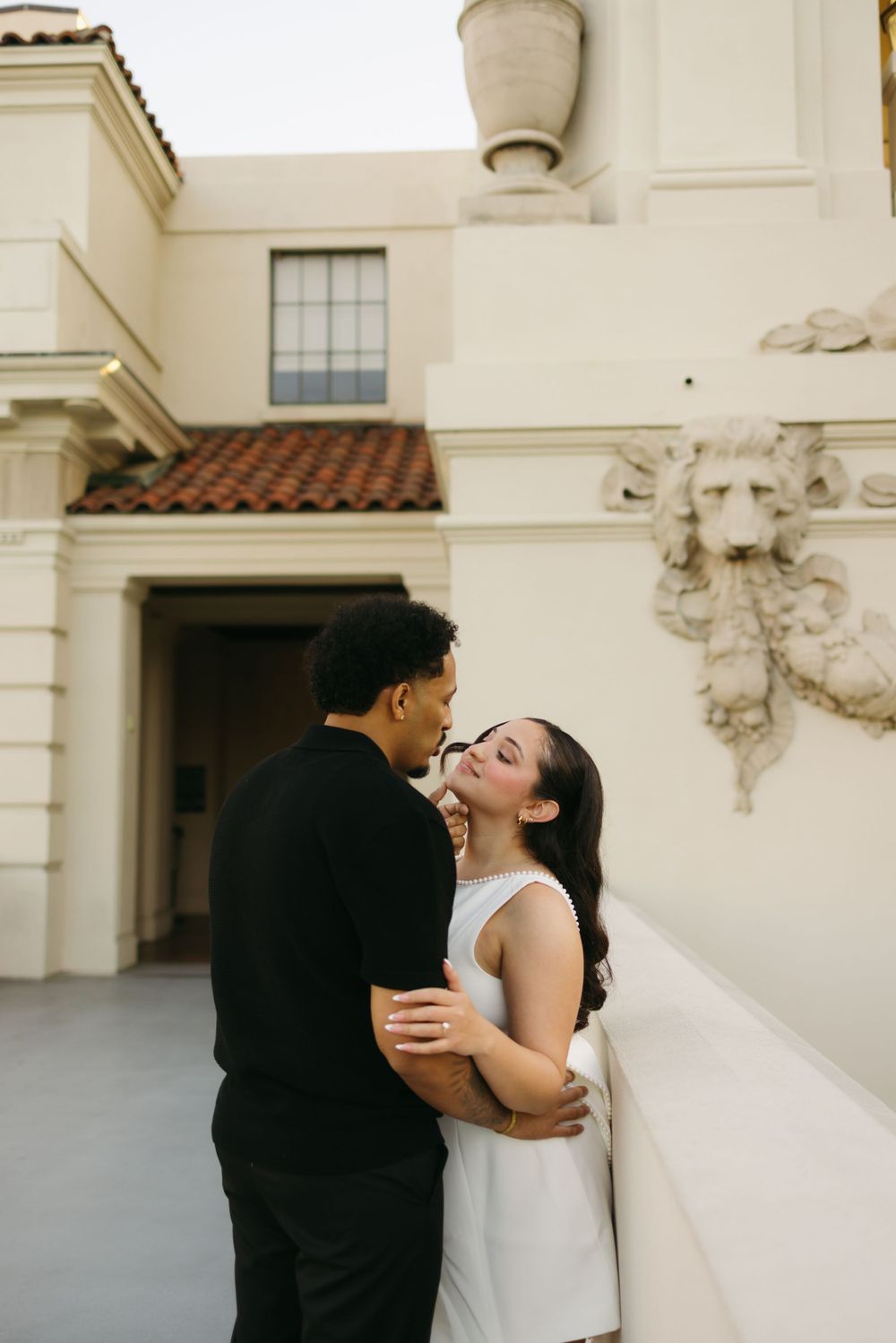 A couple shares an intimate moment against a white Mediterranean-style building with terracotta roof tiles and ornate sculpture details.