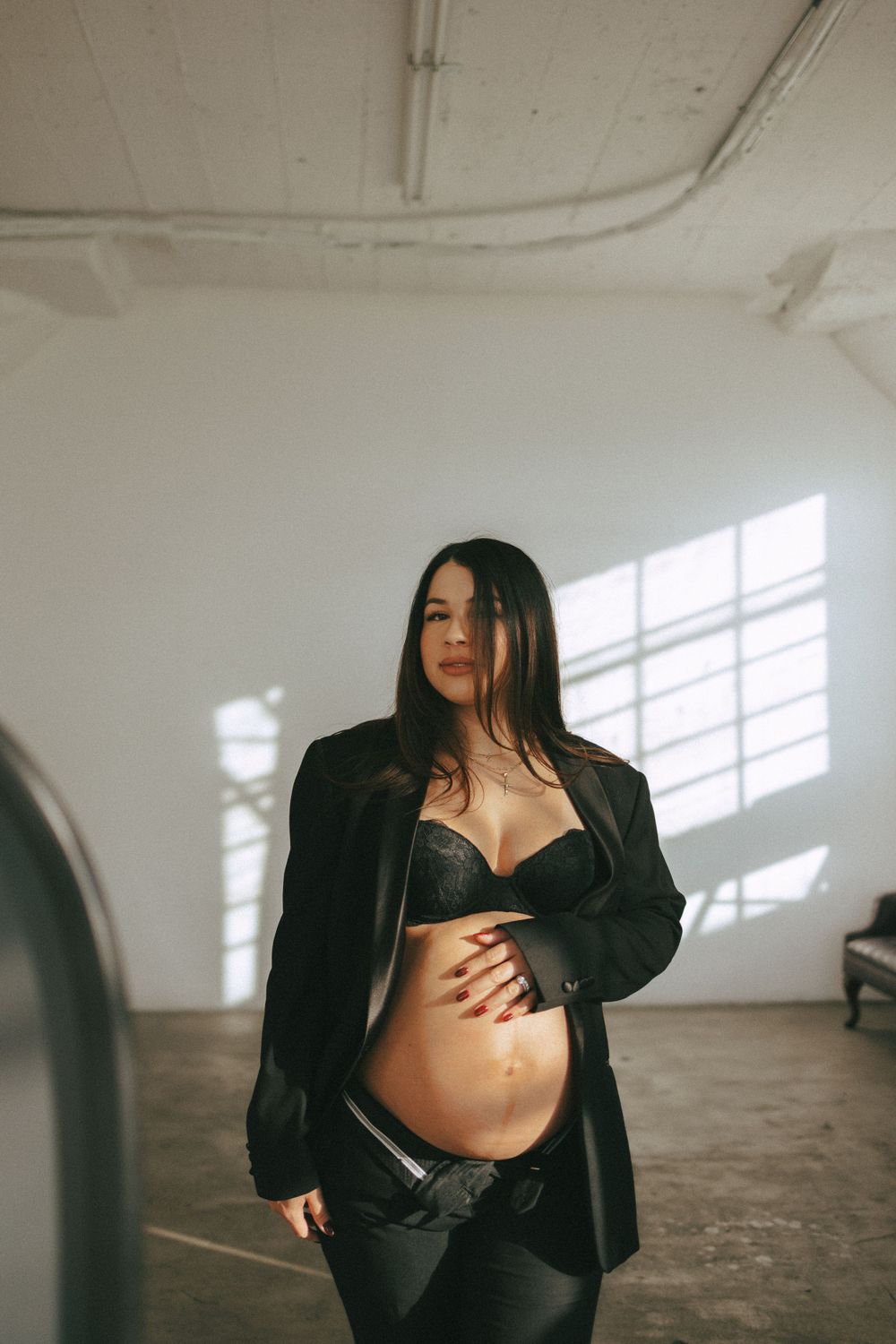 A woman poses in lingerie and an open black blazer near a window in an industrial space, casting dramatic shadows.