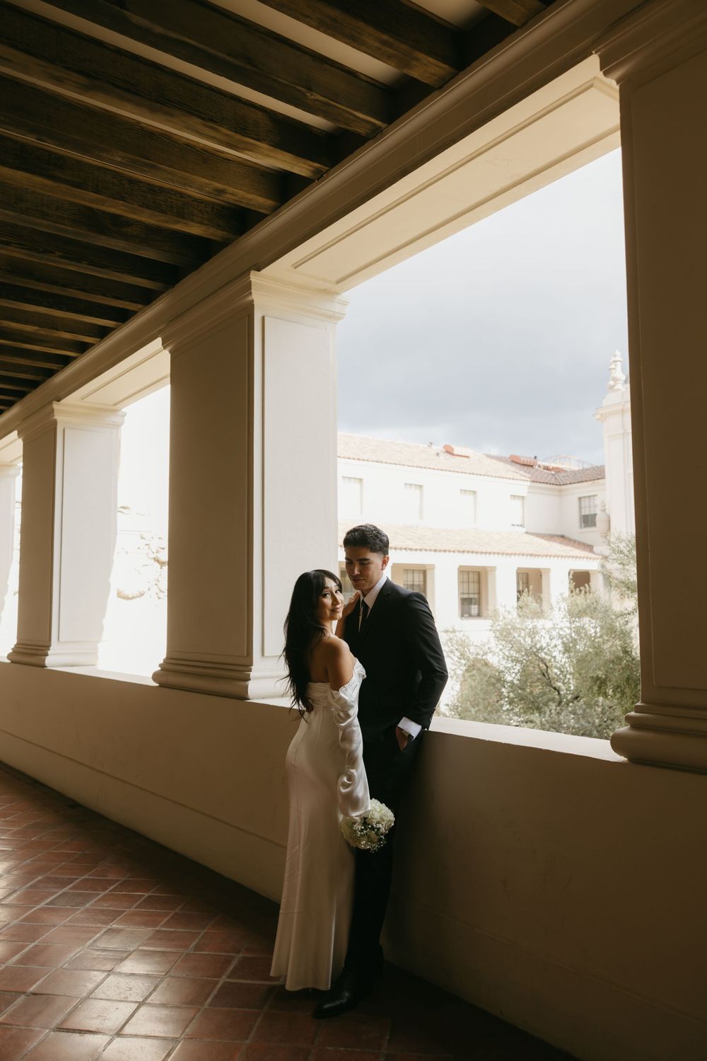 A couple in wedding attire stands together in an elegant columned hallway with terracotta floors and views of a courtyard.
