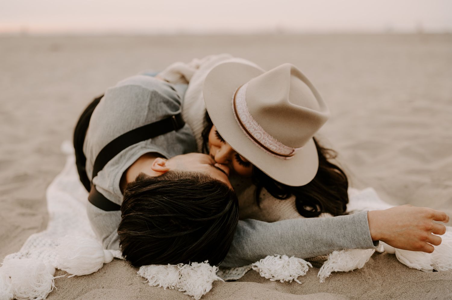 Two people in hats share intimate moment while lying on beach sand.