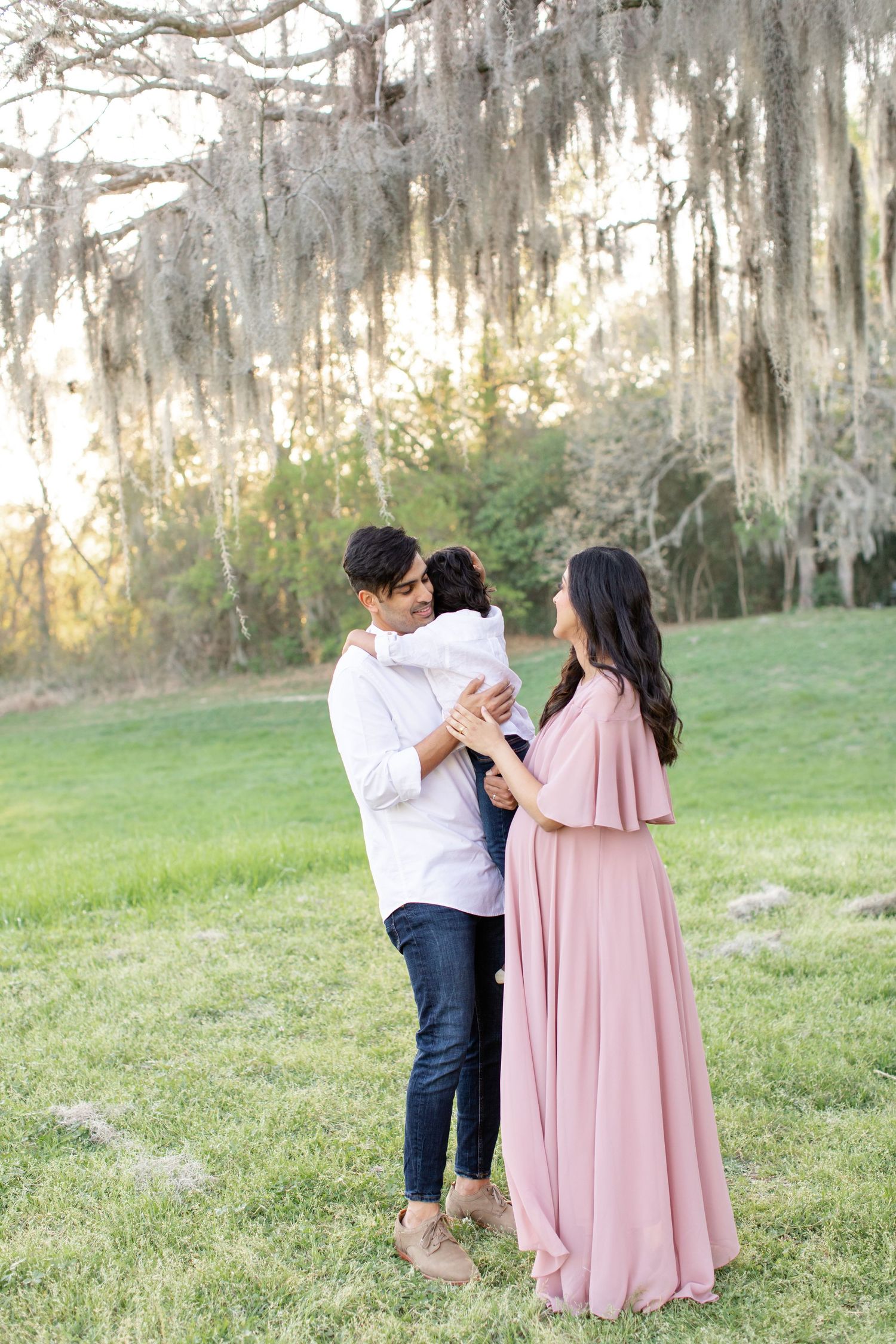 Family maternity session captures tender moments in dreamy outdoor setting with moss-draped trees.