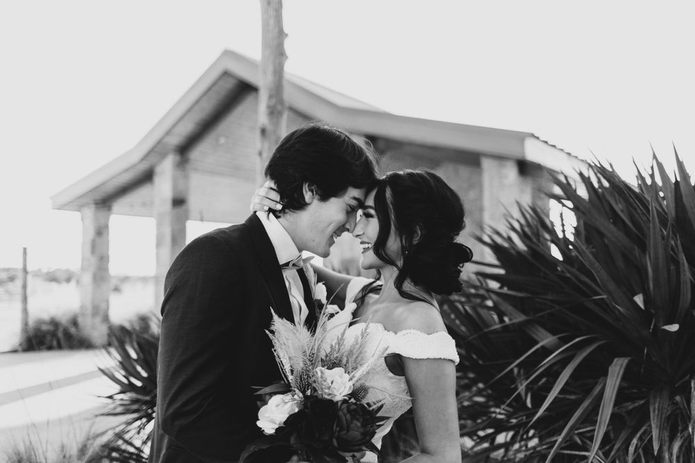 Bride and groom touch foreheads tenderly while holding a bouquet near tropical plants.