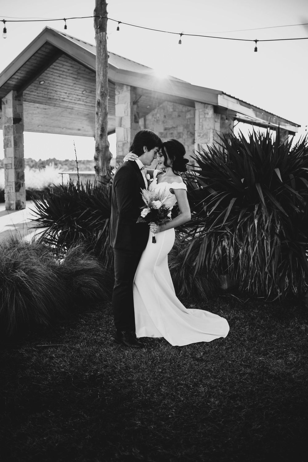 Bride and groom share a kiss outdoors near rustic building with string lights at sunset.