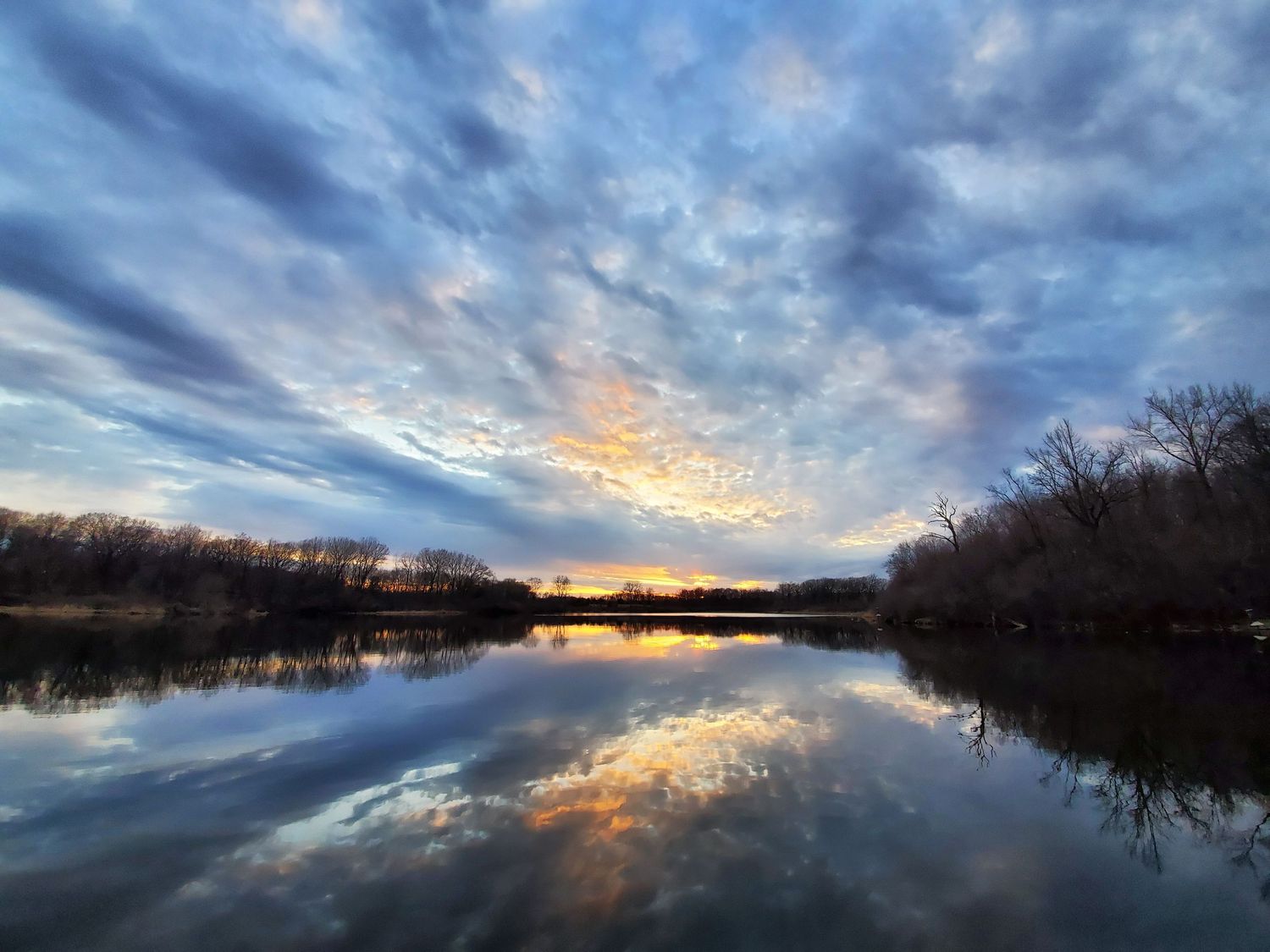Sunset reflection over a calm lake with dramatic clouds and silhouetted trees.