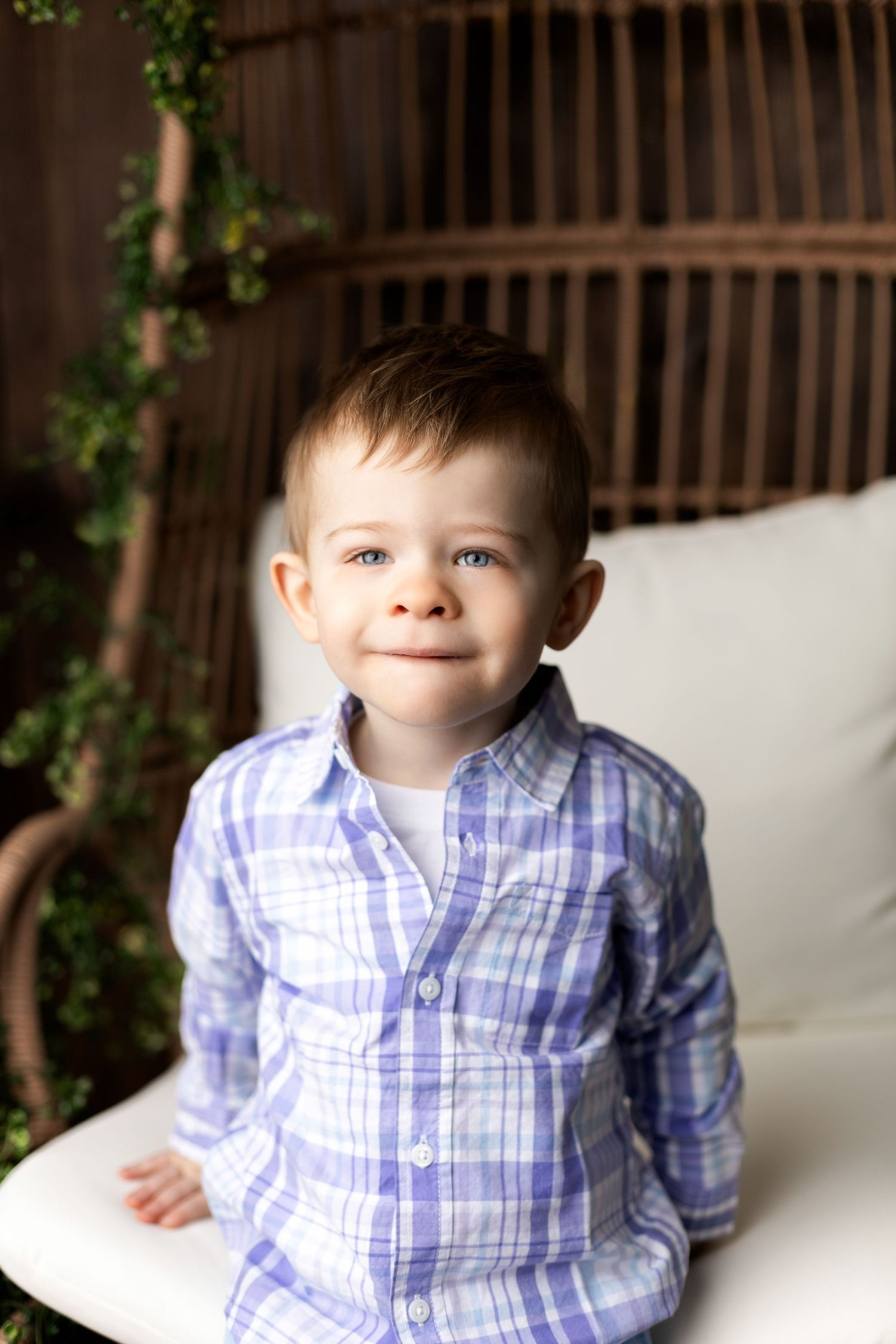 Young child in blue plaid button-down shirt sitting against outdoor backdrop.