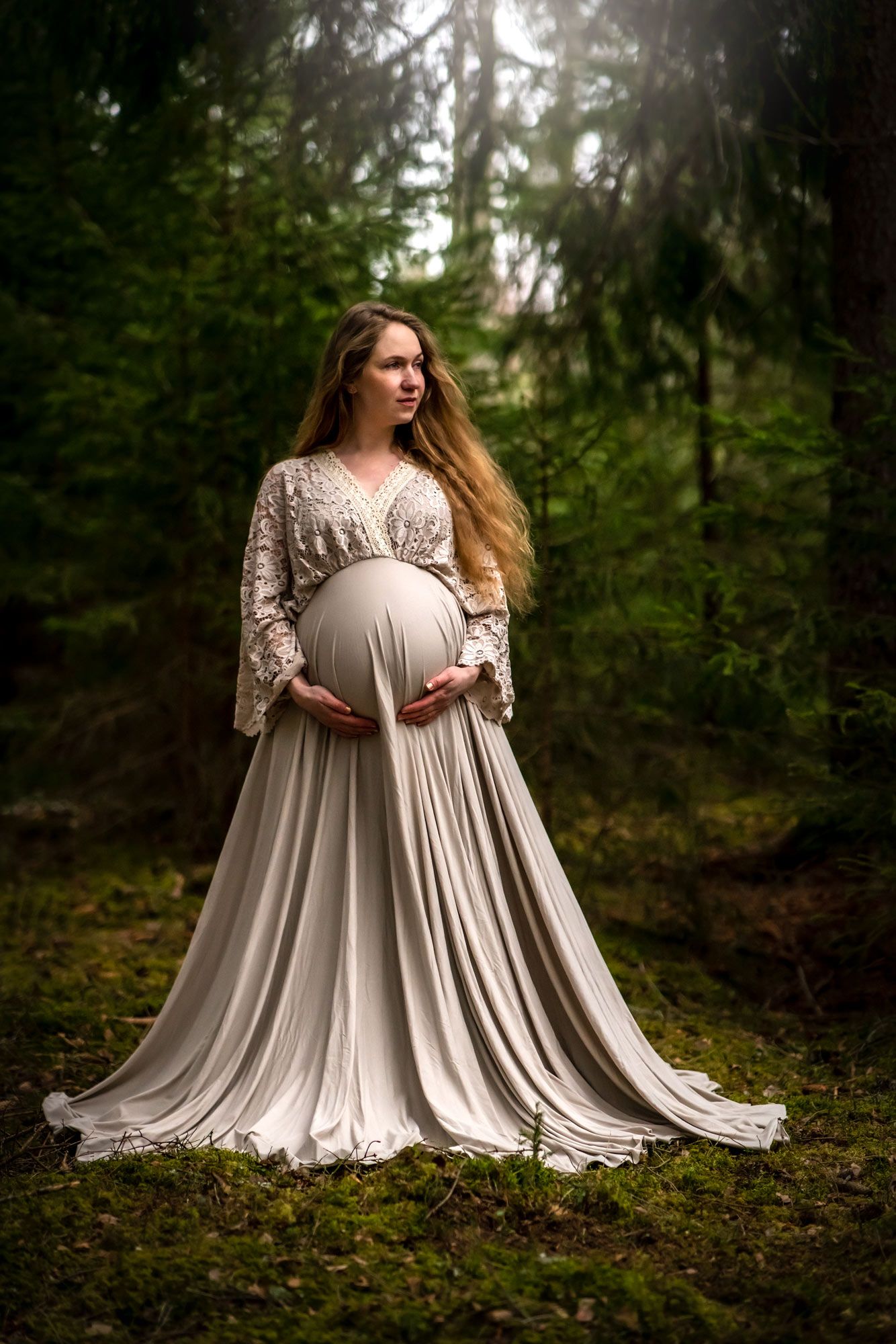 En långhårig gravid kvinna i en beige långklänning håller om sin mage. Hon står i skogen.