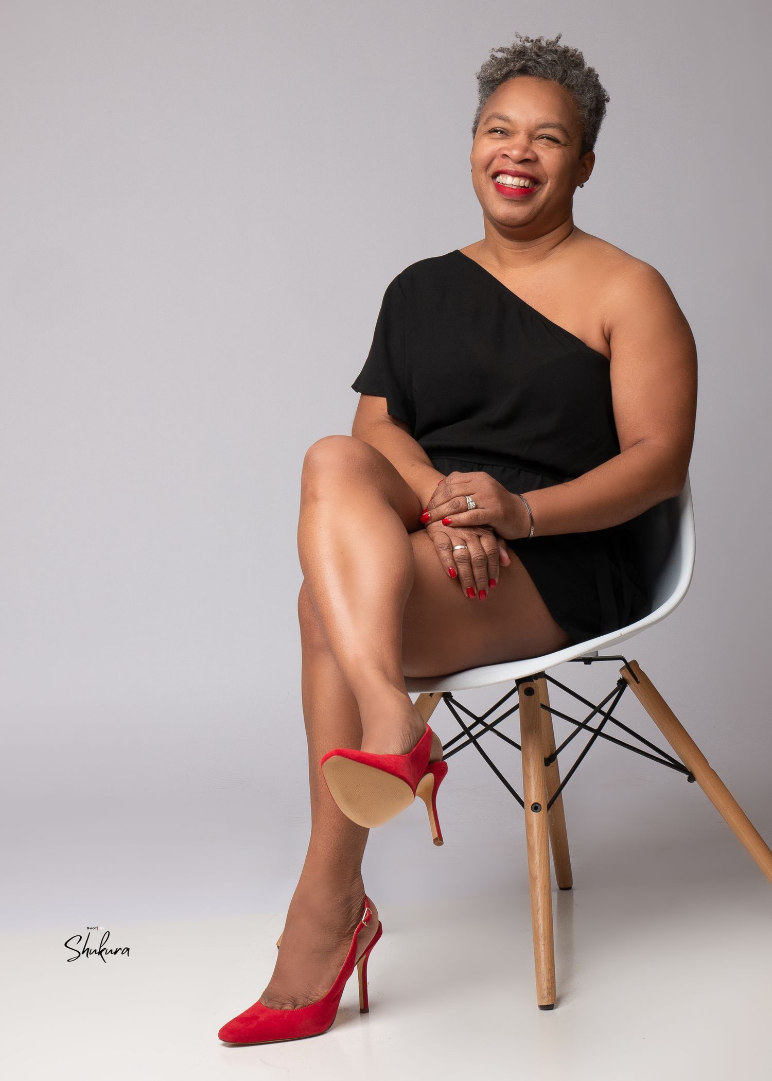 Stylish portrait featuring red heels and a black one-shoulder dress on a modern white chair.
