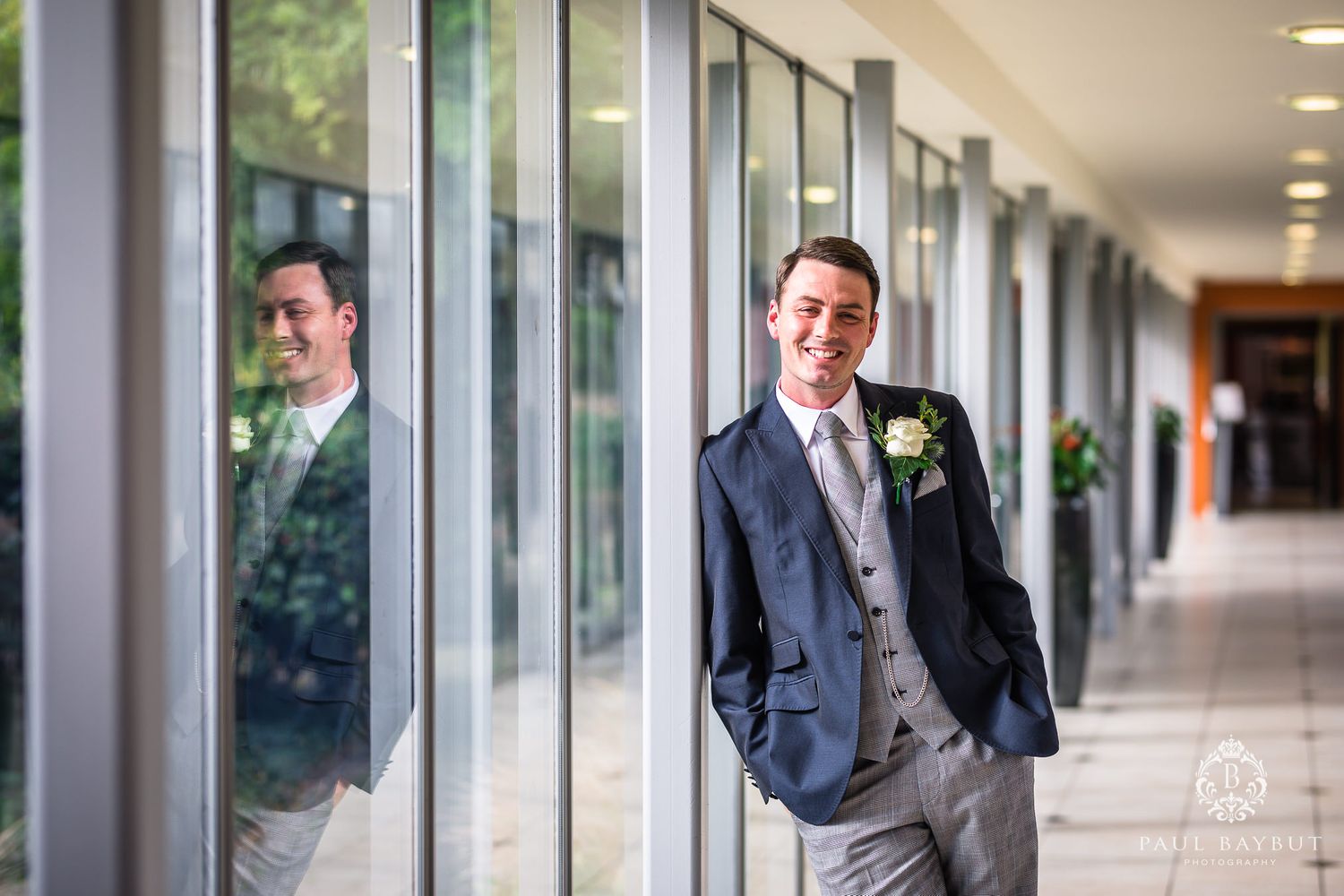 A groom in a navy suit and grey vest poses near reflective windows in an elegant hallway.