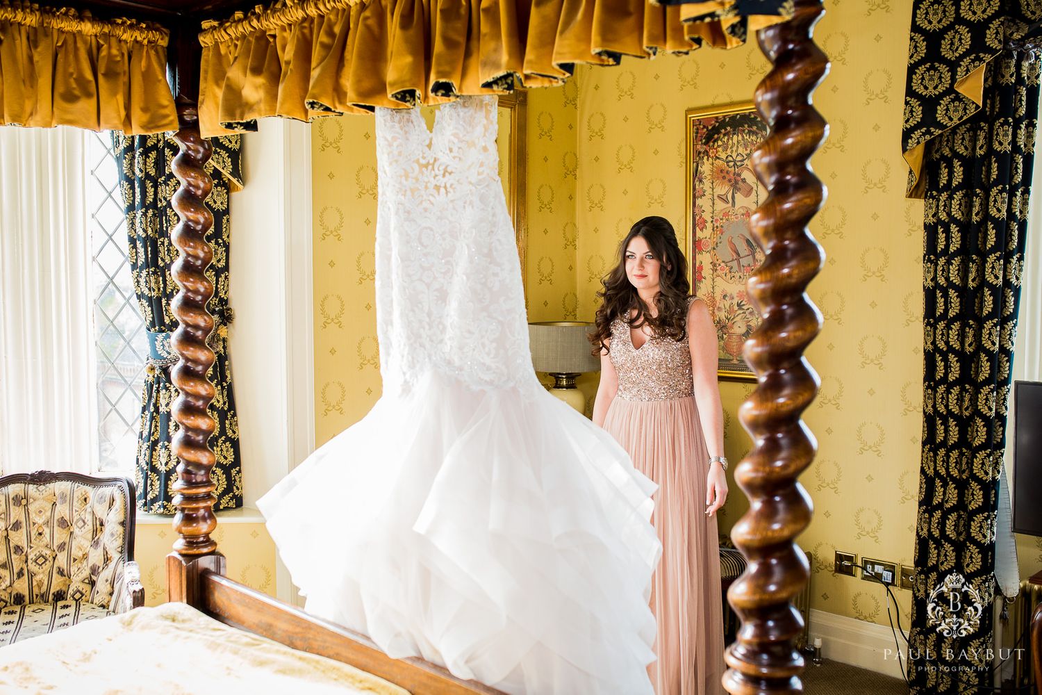 A white wedding dress hangs elegantly in a yellow room with ornate twisted wooden pillars.