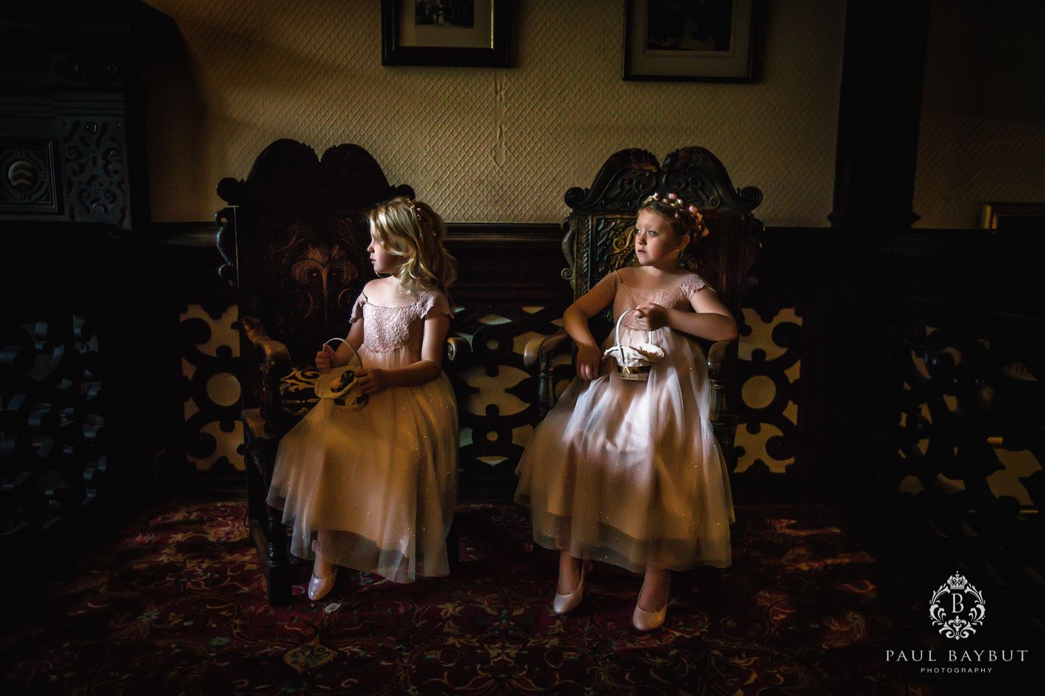 Two flower girls in matching dresses sitting on ornate wooden chairs.