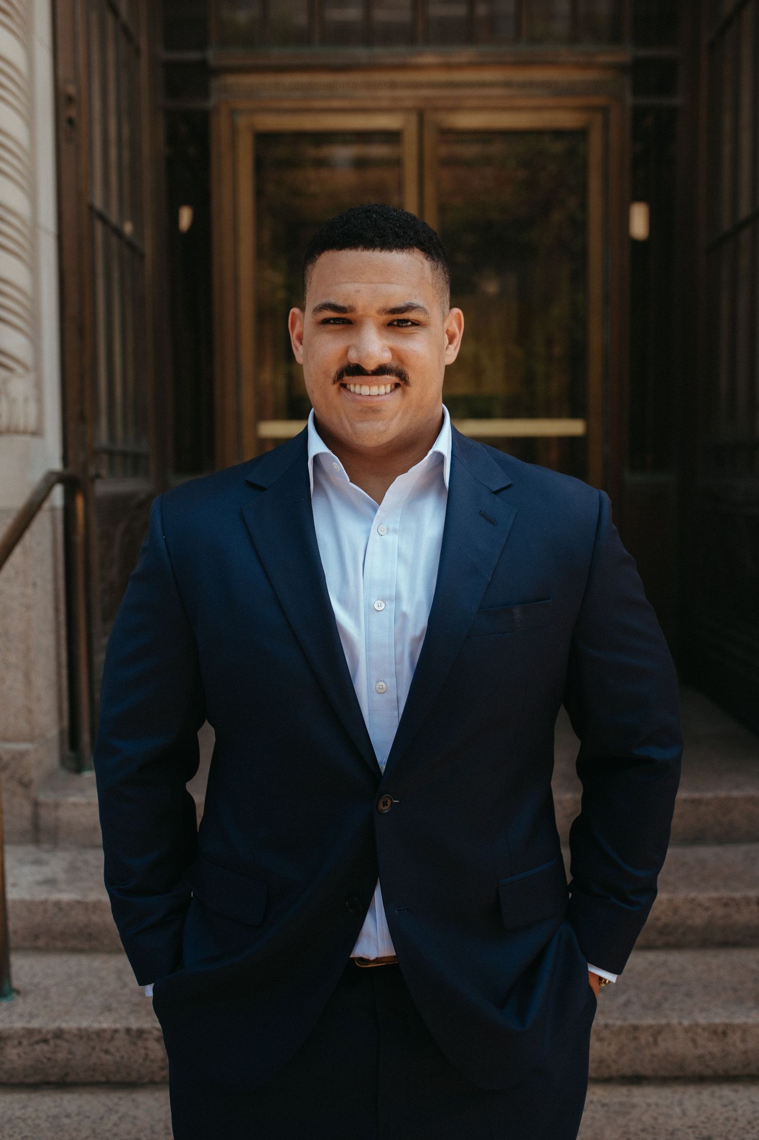 Man in navy suit and light blue shirt smiles confidently in formal portrait.