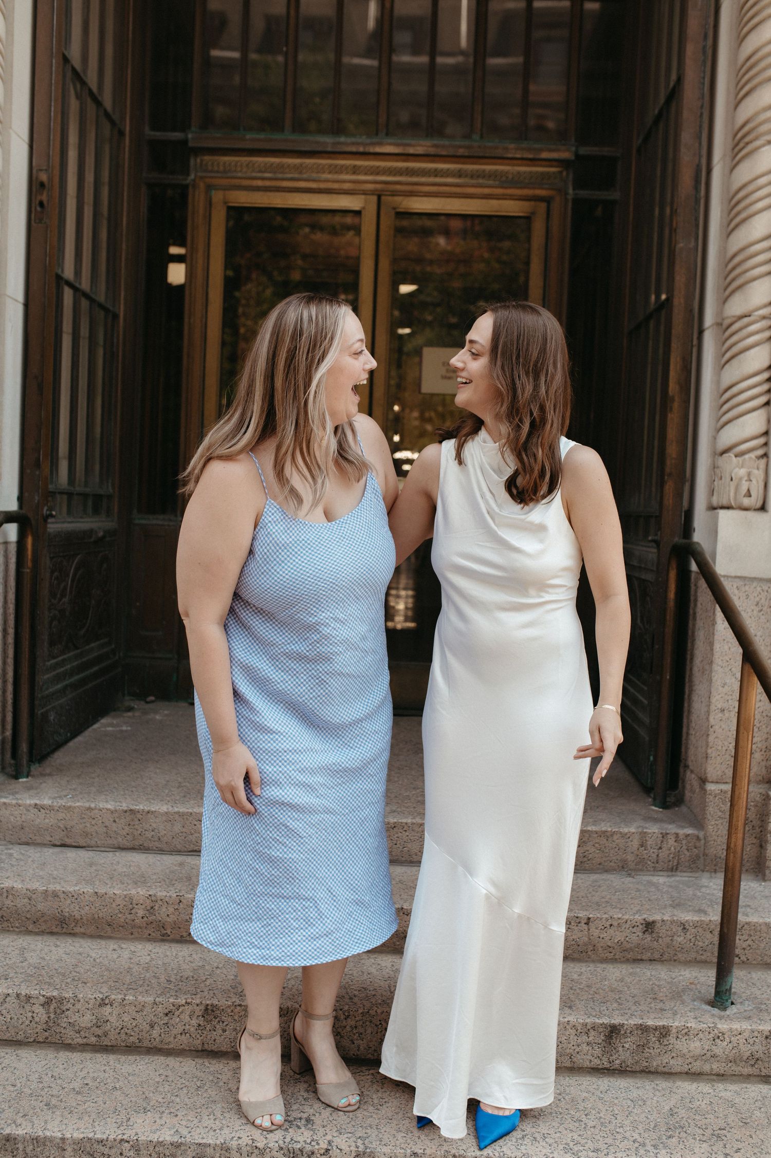 Two women in light blue and white dresses share a joyful moment on stone steps.