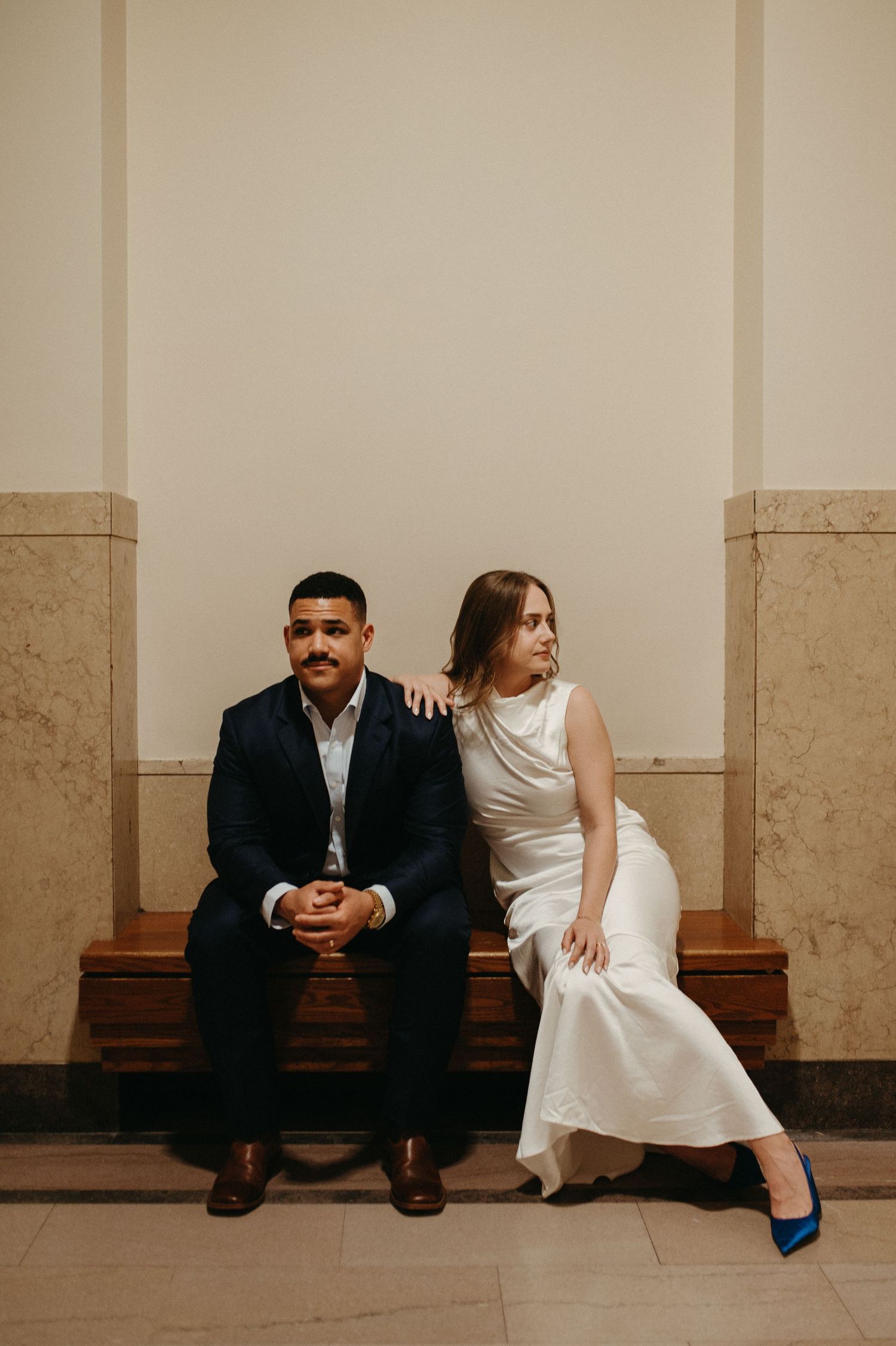 Couple in formal wear sits casually on wooden bench in classic building interior.