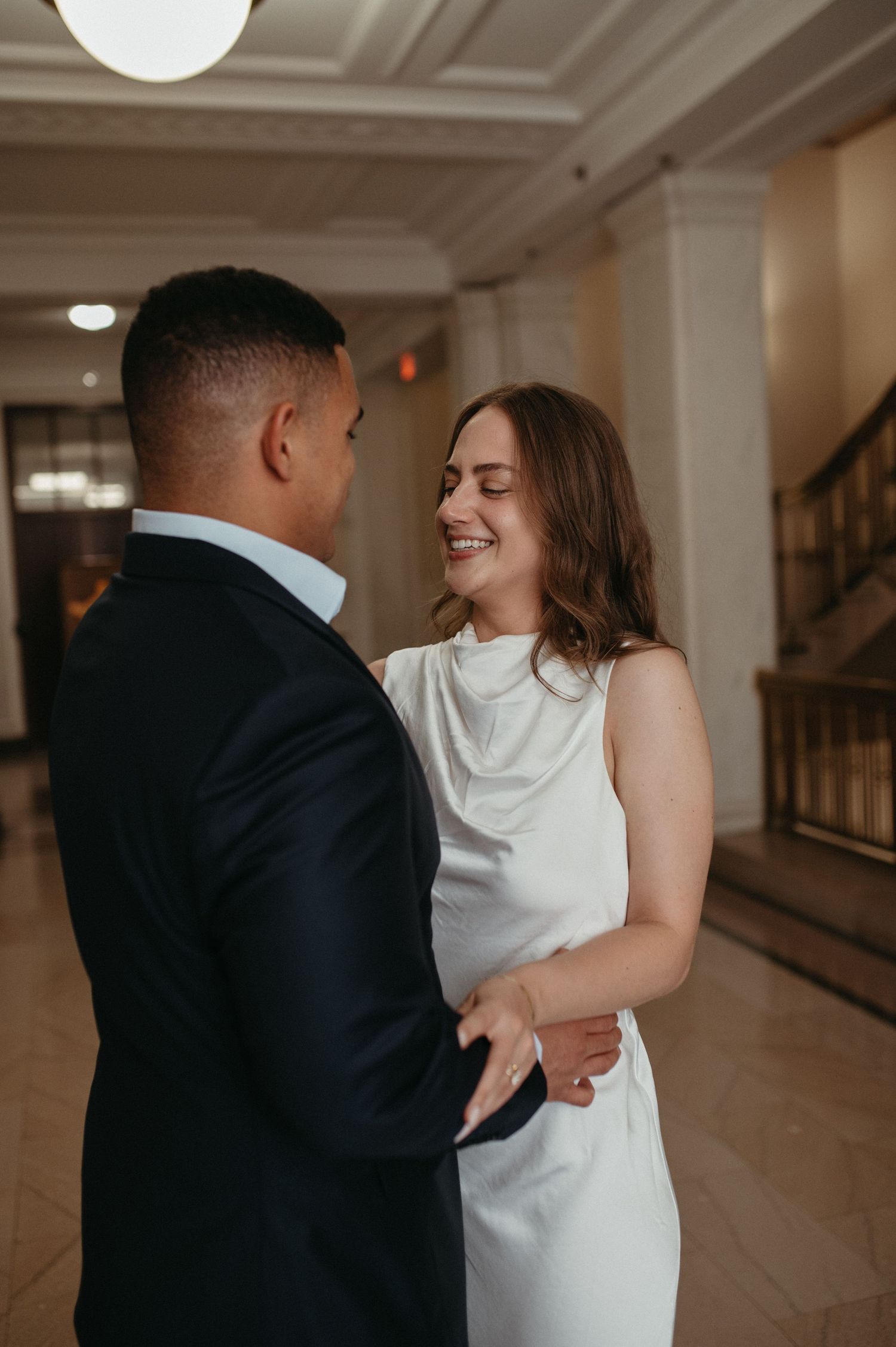 A candid moment captures a couple laughing together in formal attire inside an elegant building.
