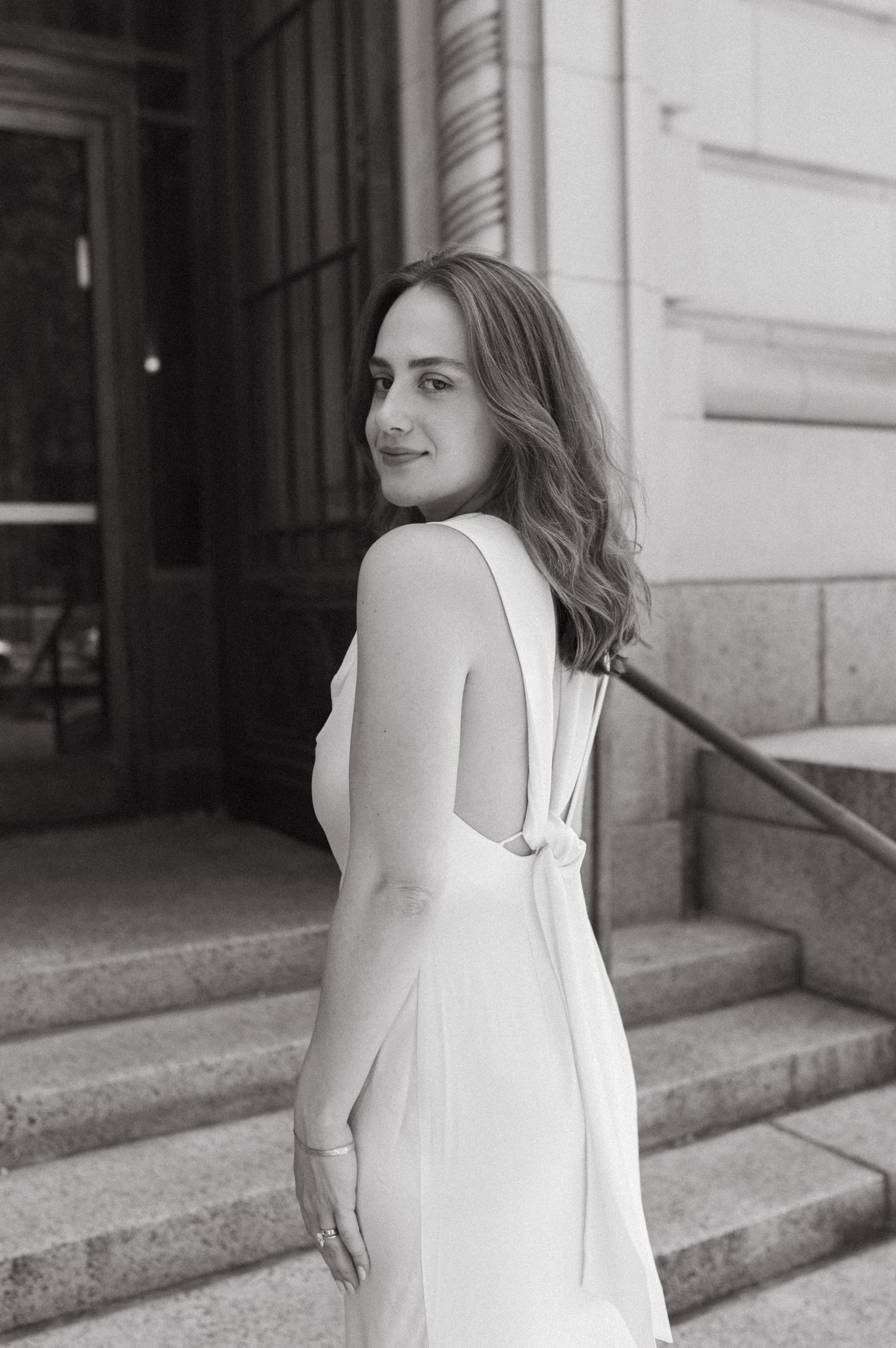 Woman in white formal dress poses on stone steps showing elegant back detail.